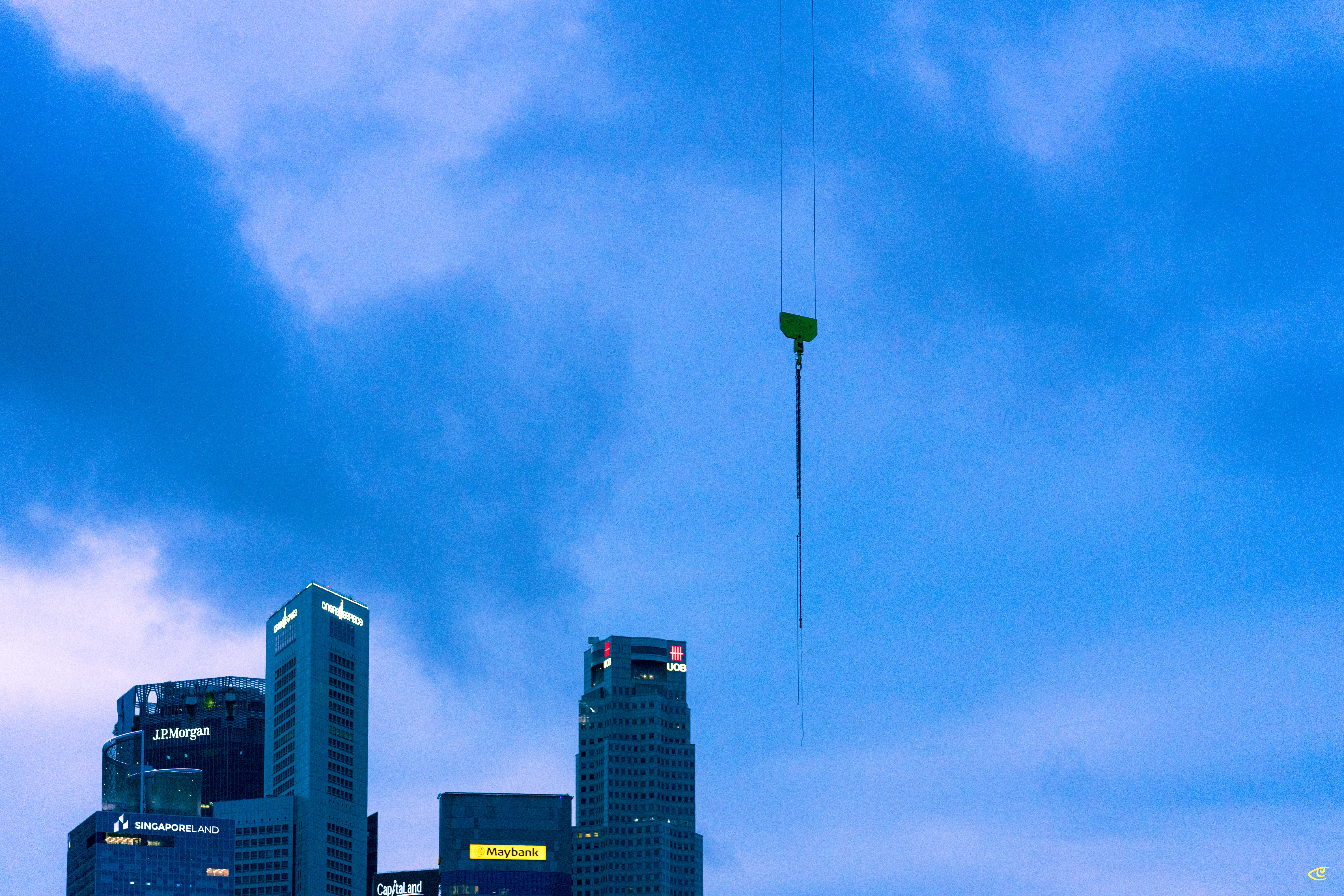 Suspended crane hook hanging from cables against a cloudy blue sky above a city skyline with several high-rise buildings and illuminated signs near the bottom of the frame.