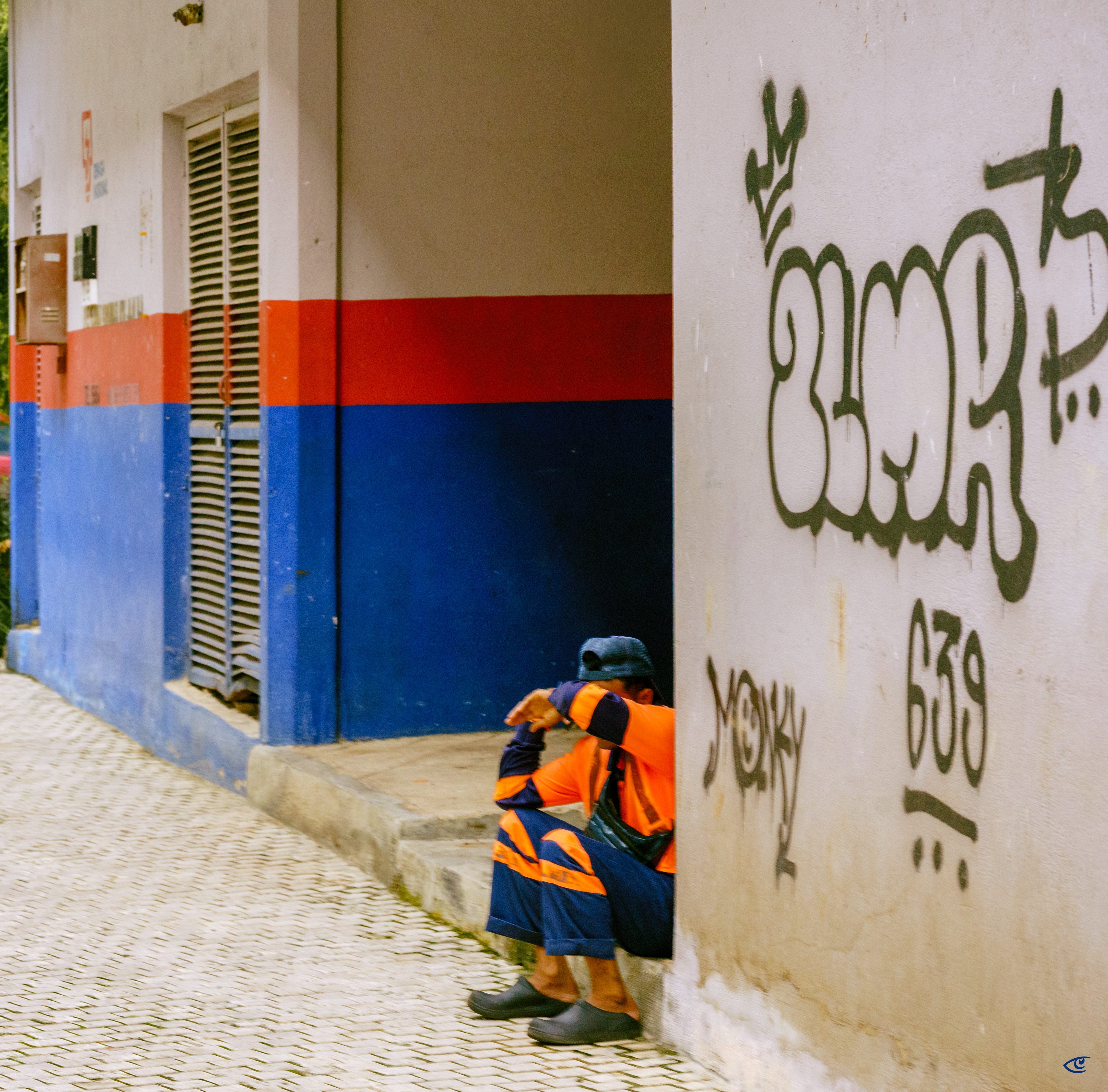 Person in an orange-and-navy reflective work uniform sits on a curb with head resting on crossed arms, partially hidden by a white wall covered in green graffiti; cobblestone sidewalk and a building facade painted blue with a red stripe and a metal shuttered doorway in the background.