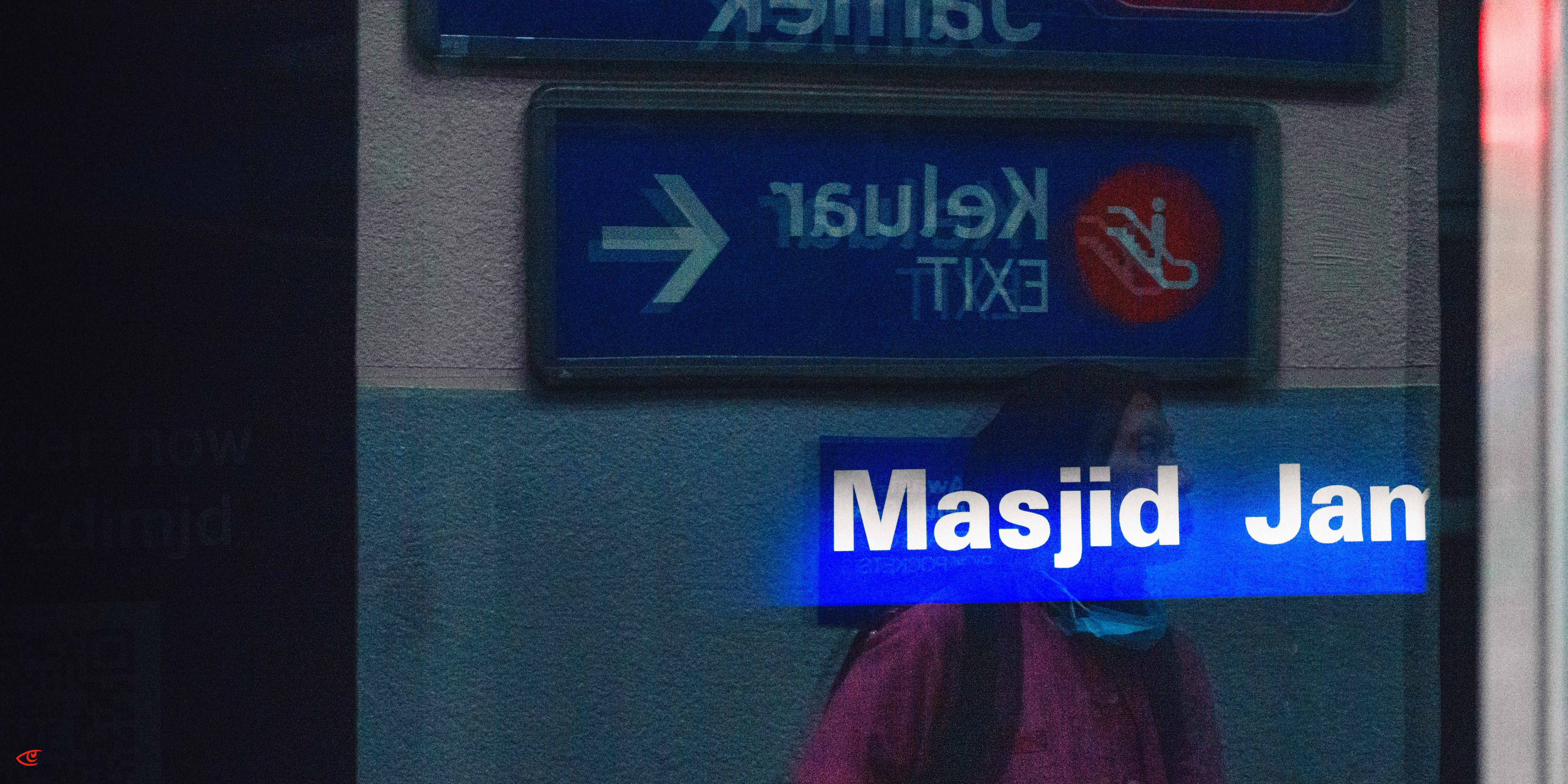 Blurred view through glass of a woman in a pink top wearing a face mask walking past a blue sign reading “Masjid Jan,” with a reversed “Keluar EXIT” sign and right-pointing arrow above.
