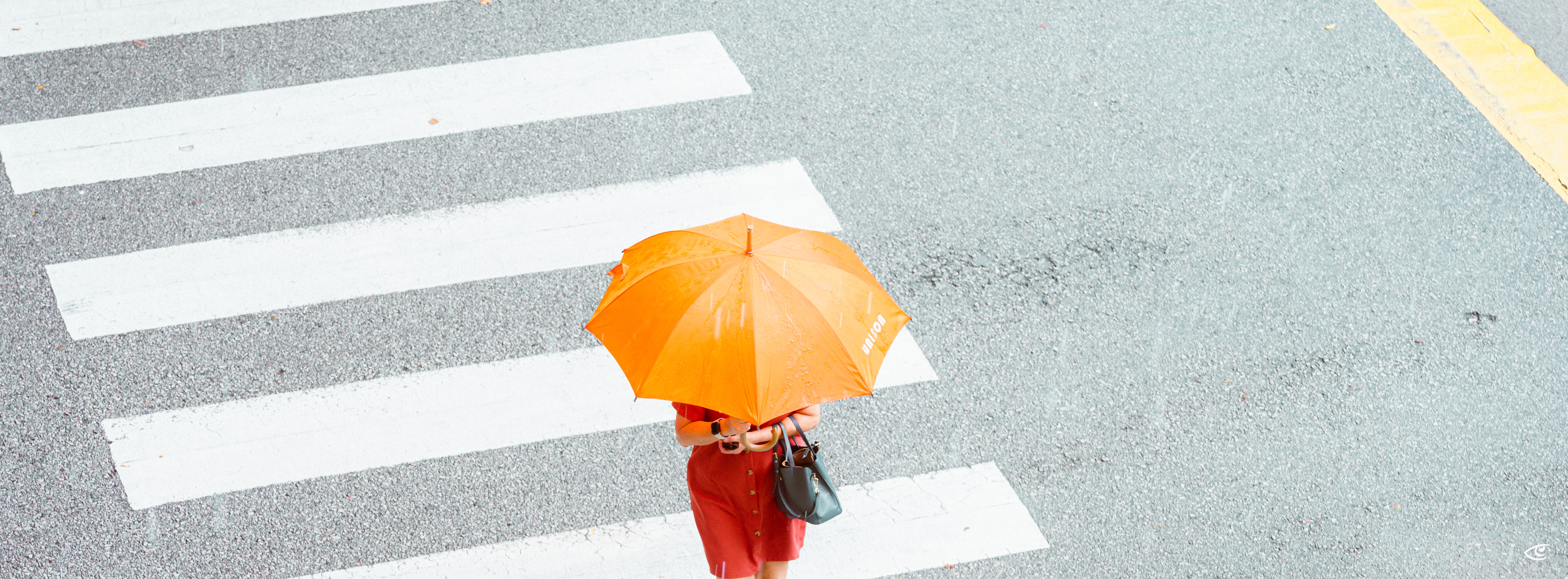 Overhead view of a pedestrian holding an orange umbrella and wearing a red outfit while crossing a street on a white-striped crosswalk.