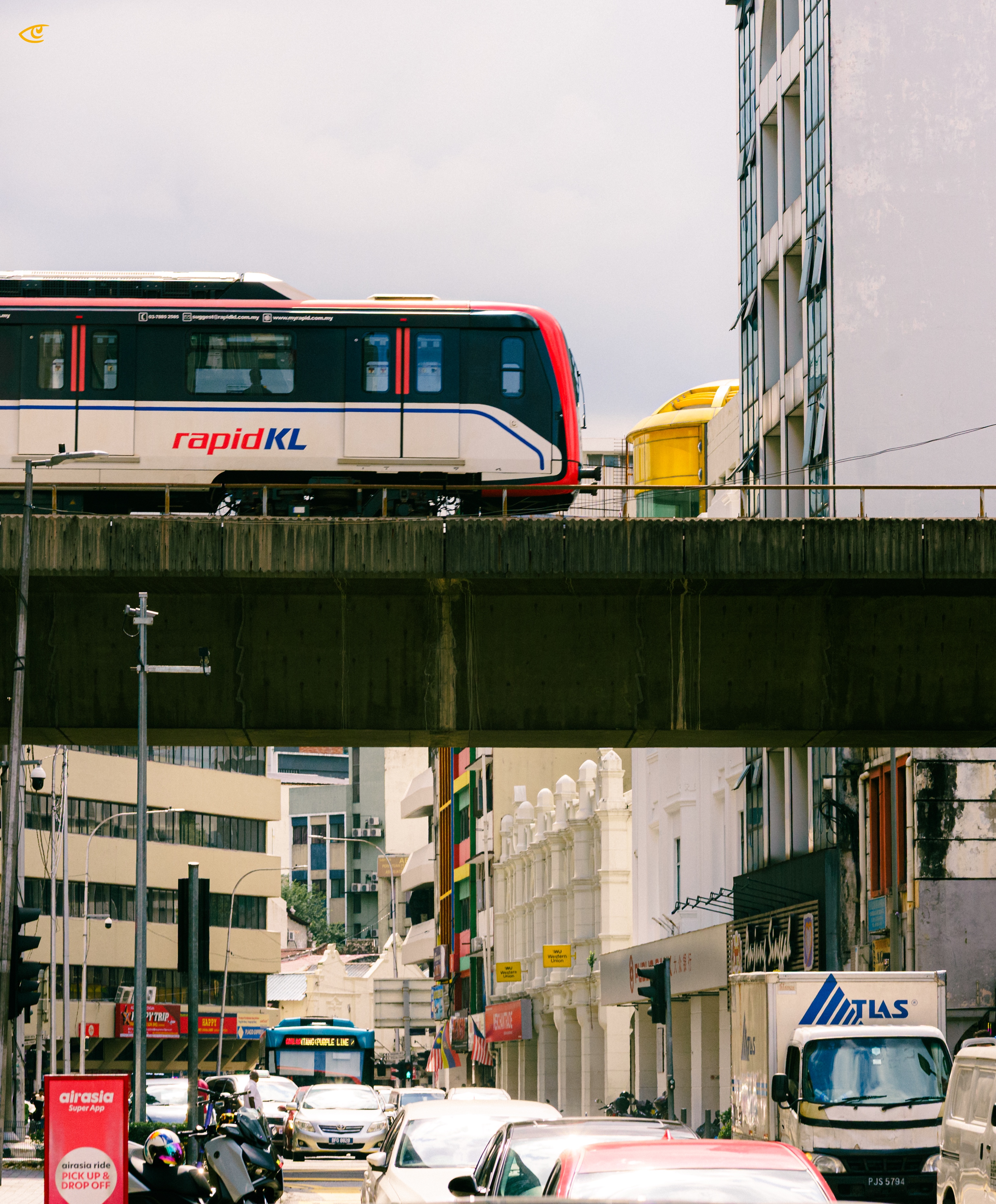 Elevated RapidKL train crossing a concrete viaduct above a busy city street with cars, motorcycles, and a delivery truck, flanked by mid-rise buildings under an overcast sky.