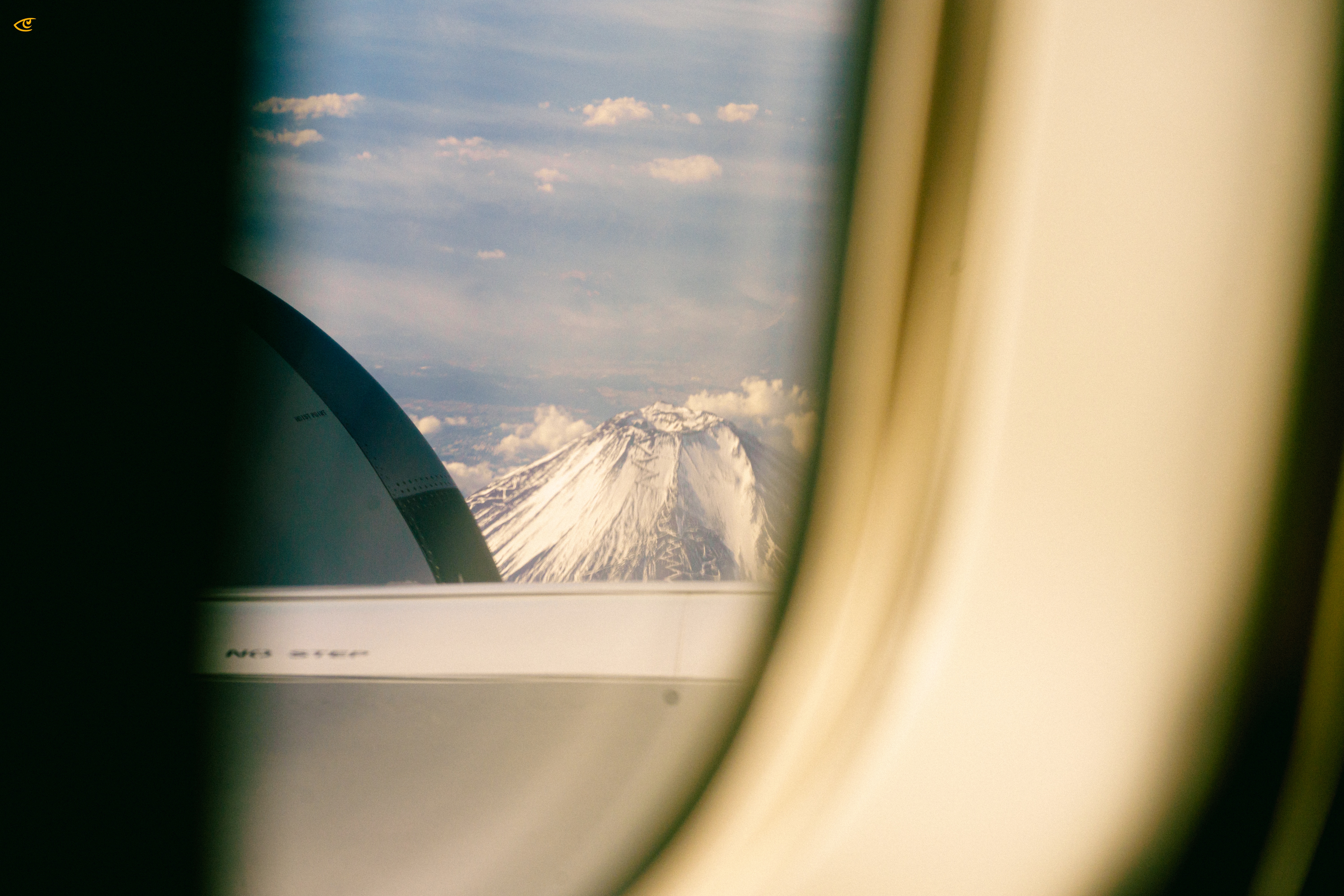 View through an airplane window showing the wing and engine in the foreground and a snow-covered mountain peak rising above clouds under a blue sky.