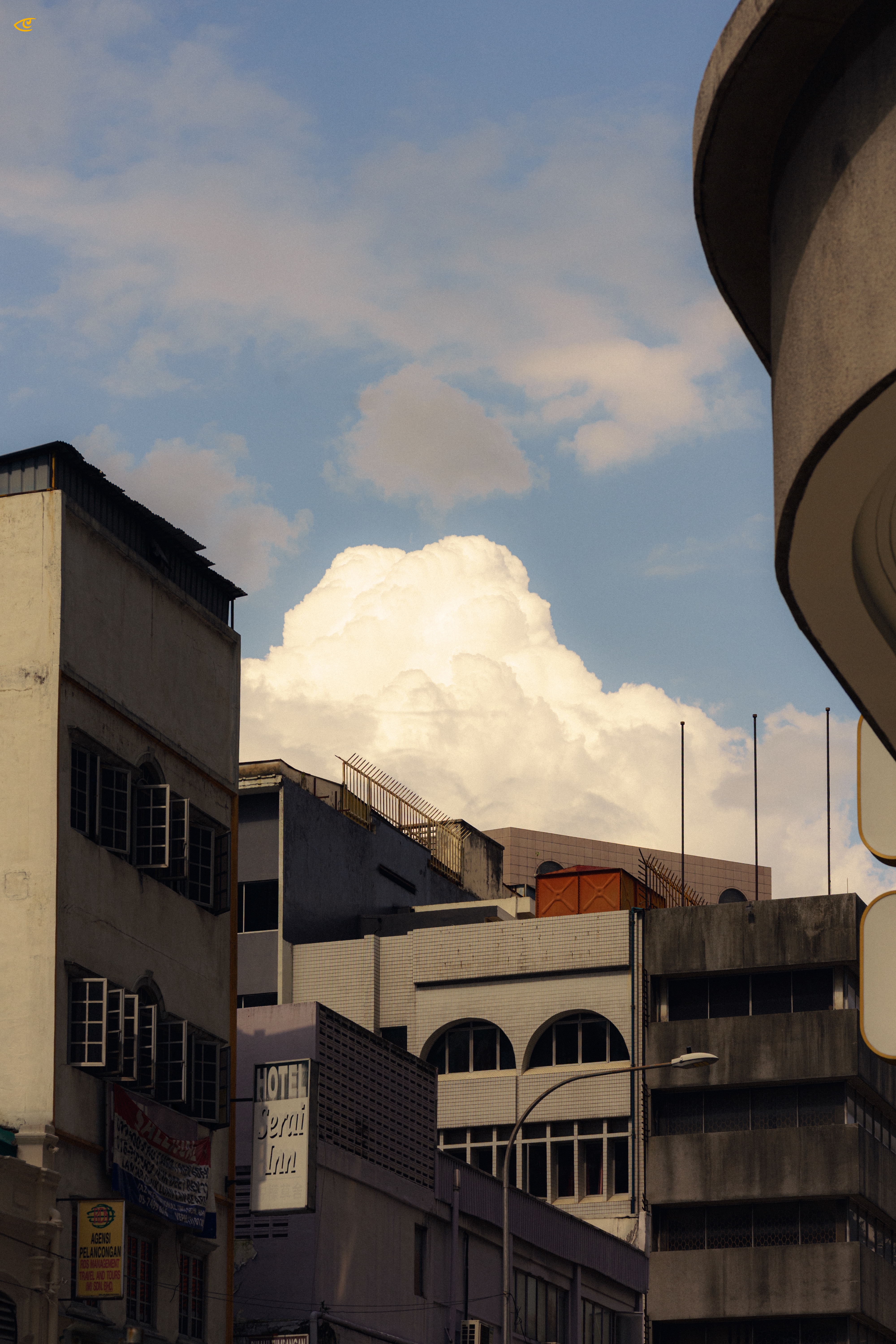 Low-angle view of older urban buildings with weathered facades and rooftop railings, a partially visible “HOTEL” sign on the left, and a streetlight in the foreground; a large white cumulus cloud rises behind the rooftops against a blue sky, with a curved building edge framing the right side.
