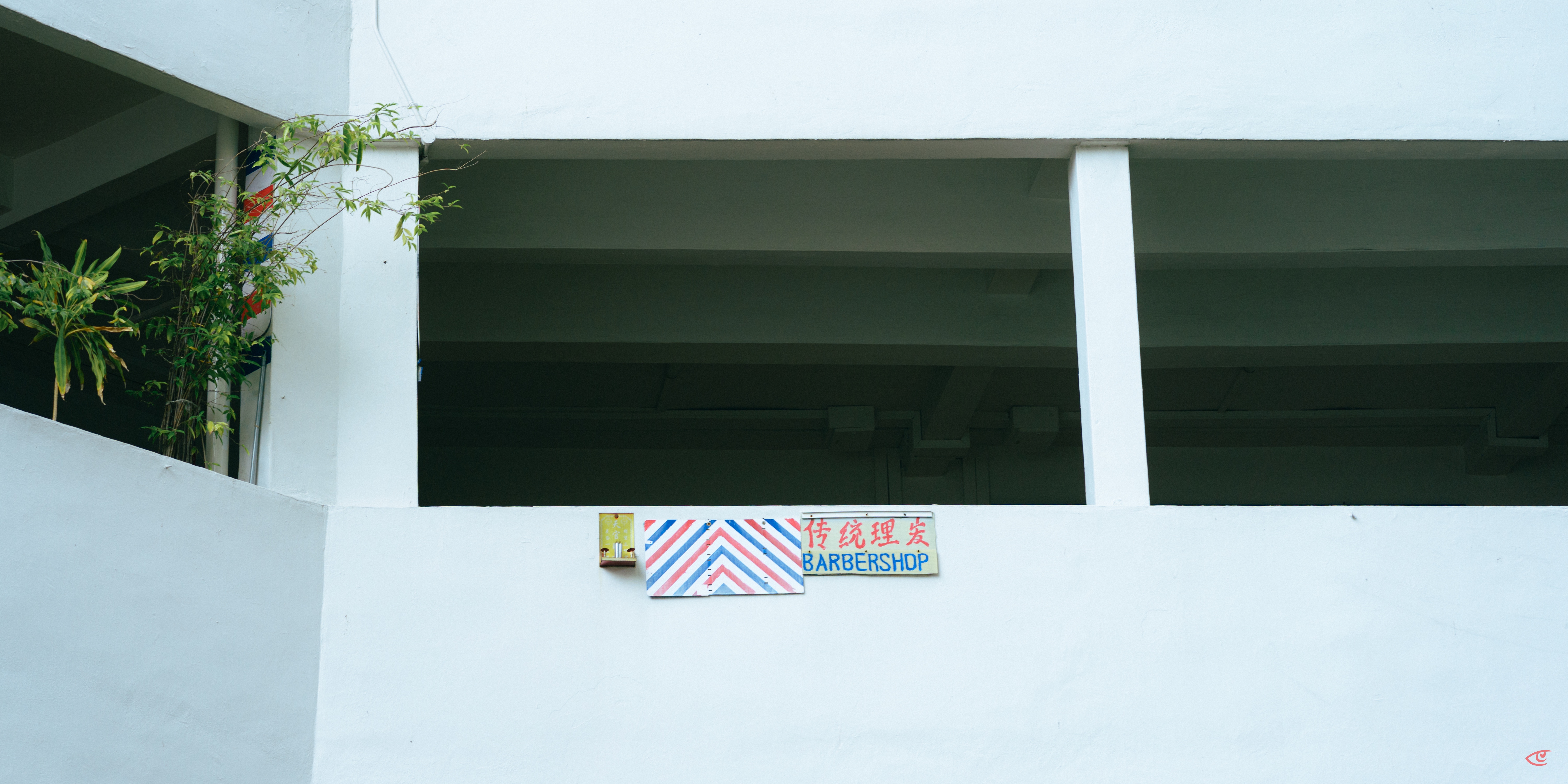Upper level of a white concrete building with open rectangular bays; a small sign reading “BARBERSHOP” with red-and-blue diagonal stripes and Chinese characters is mounted at center, with green plants hanging from the left corner.