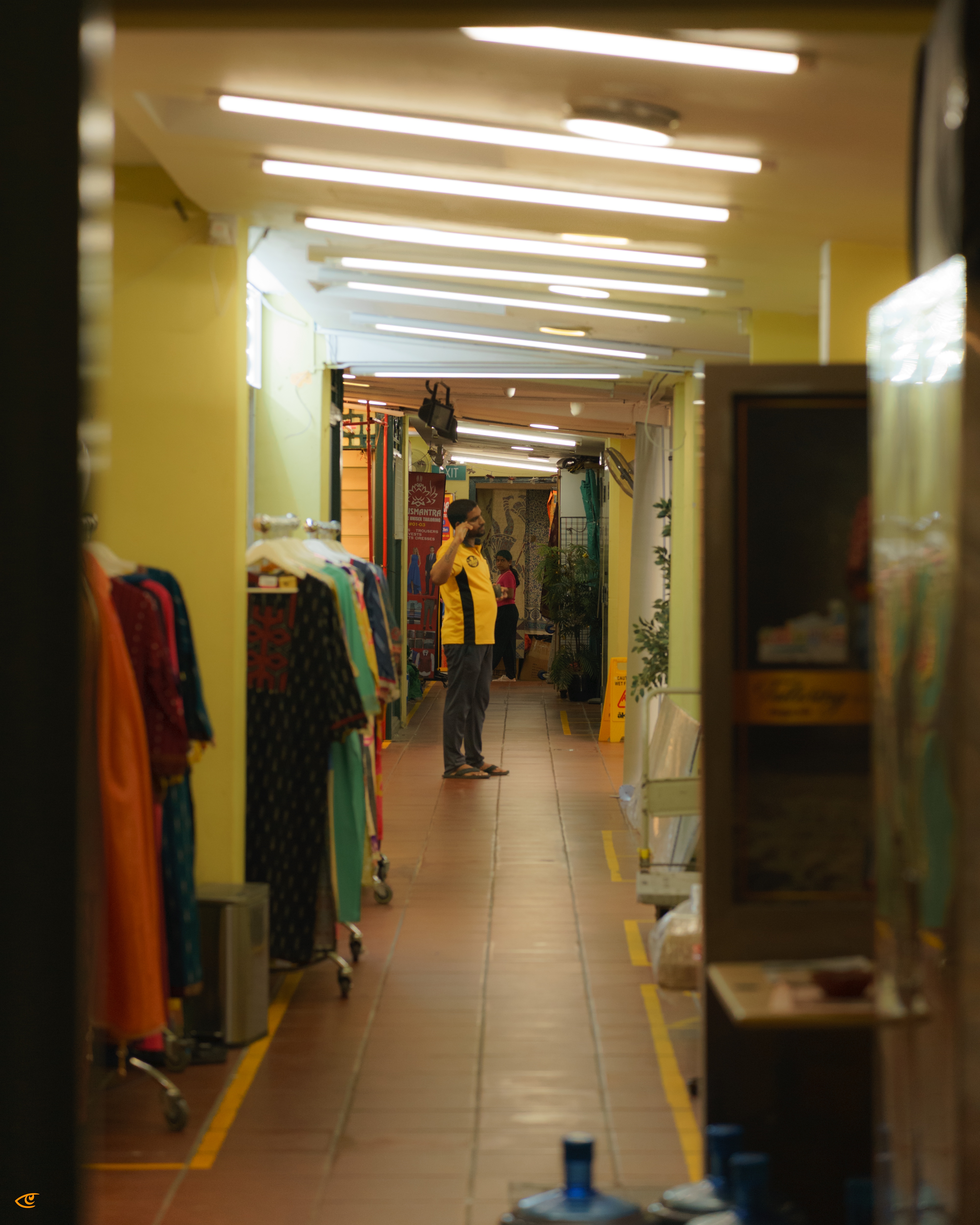 Long indoor corridor with fluorescent ceiling lights, clothing racks along the left side, and a man in a yellow-and-black shirt standing near the center; blurred shelves and large water jugs in the foreground on the right.