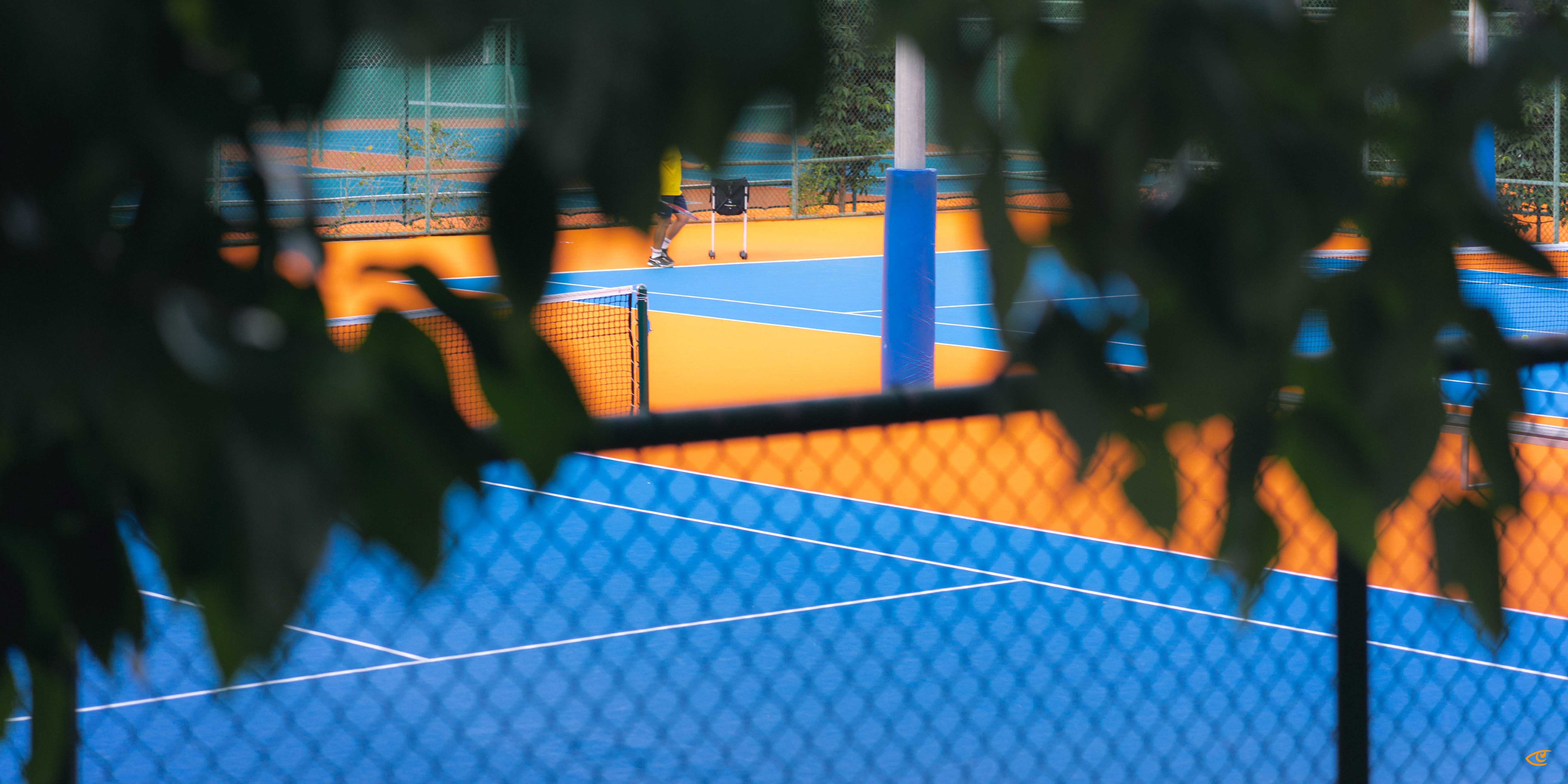 Blue and orange tennis courts viewed through a chain-link fence and out-of-focus tree leaves in the foreground, with a small figure near the far baseline and court net and posts visible.