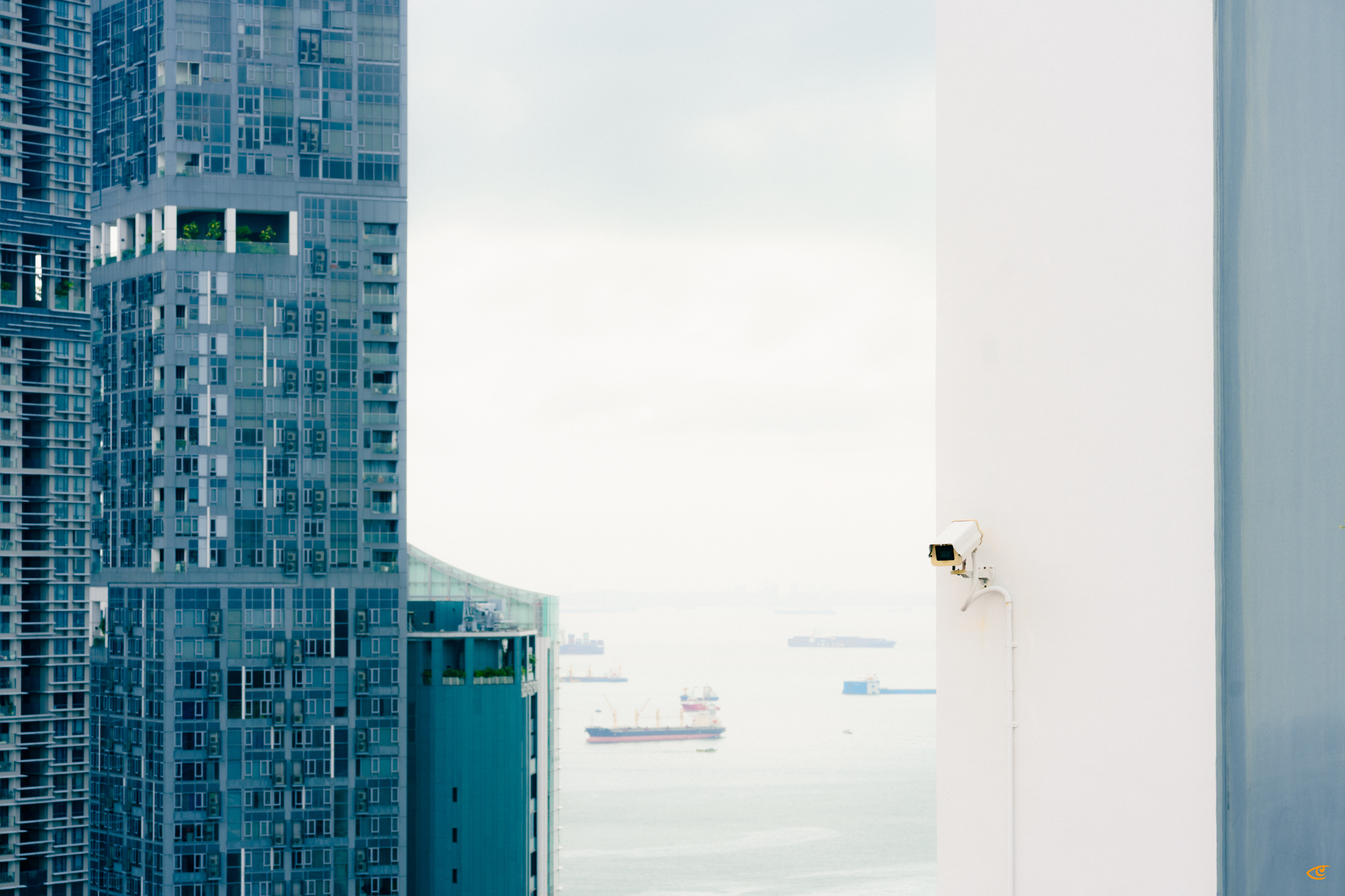 Glass high-rise building on the left and a plain white building wall on the right with a mounted security camera; distant ocean view with several cargo ships visible in the gap between the buildings under a cloudy sky.