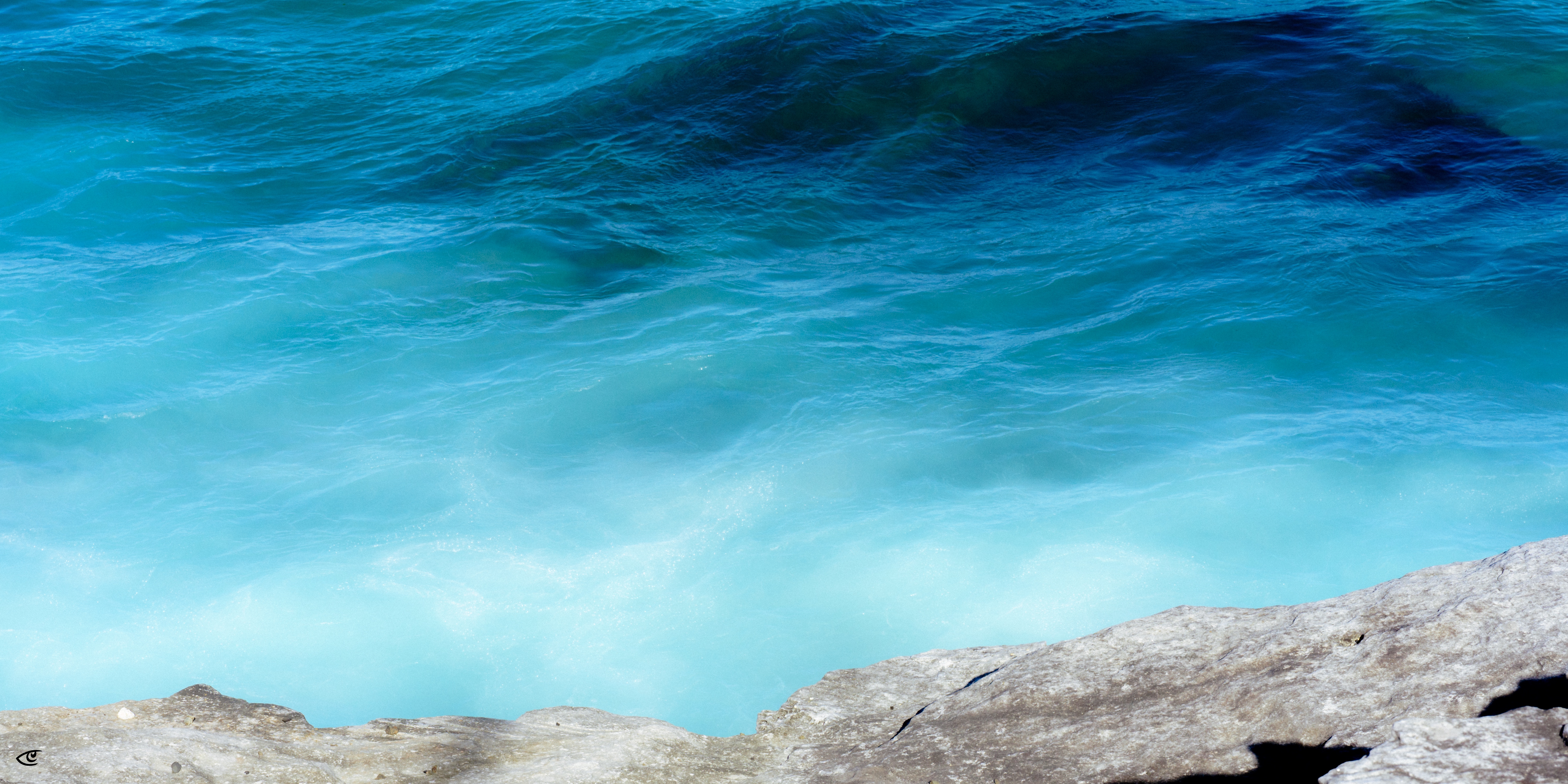 Turquoise ocean water with a darker blue wave near the top, viewed from above, with a light gray rocky shoreline along the bottom edge.