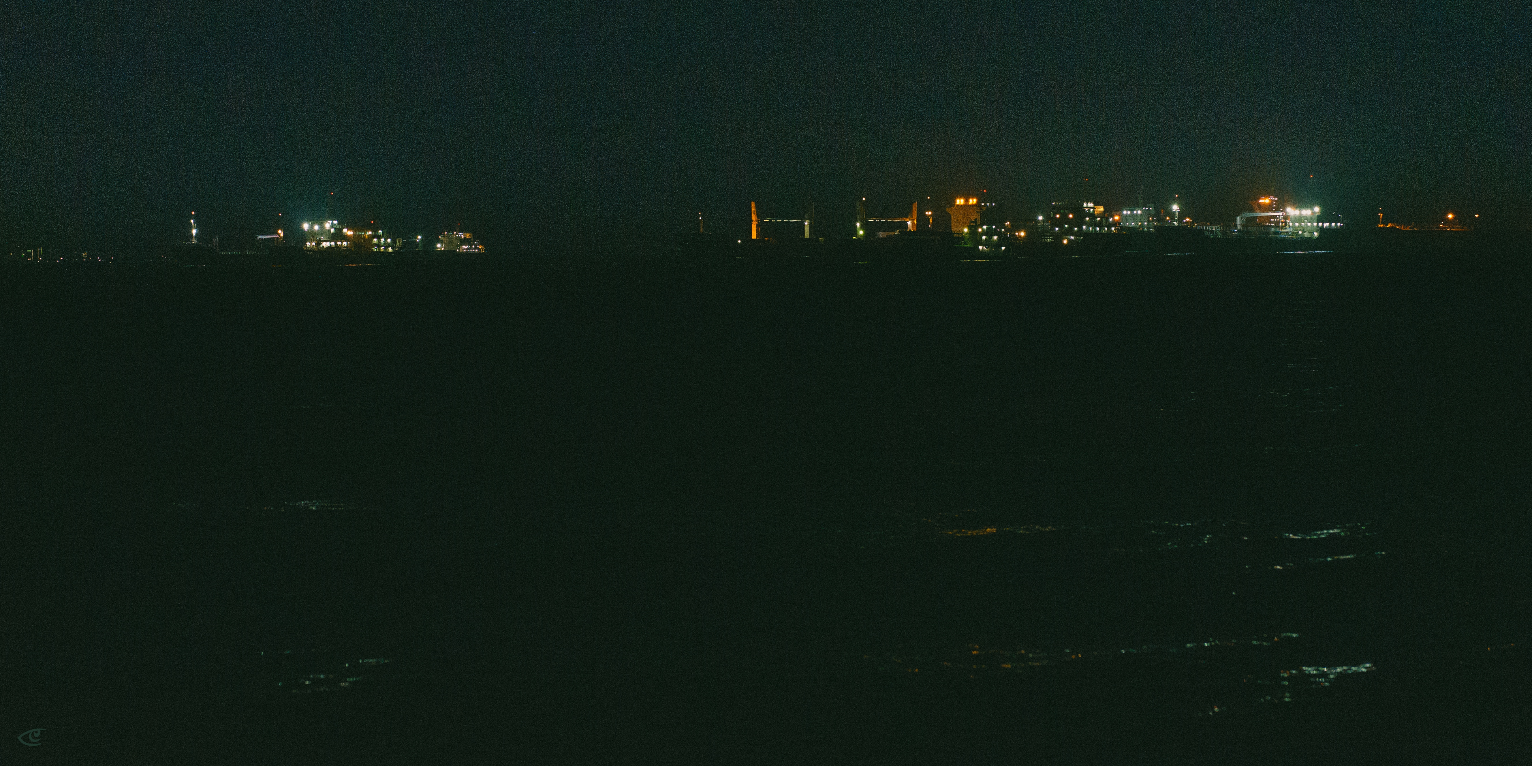 Distant shoreline at night with scattered city and industrial lights along the horizon above dark water, viewed from across the sea.