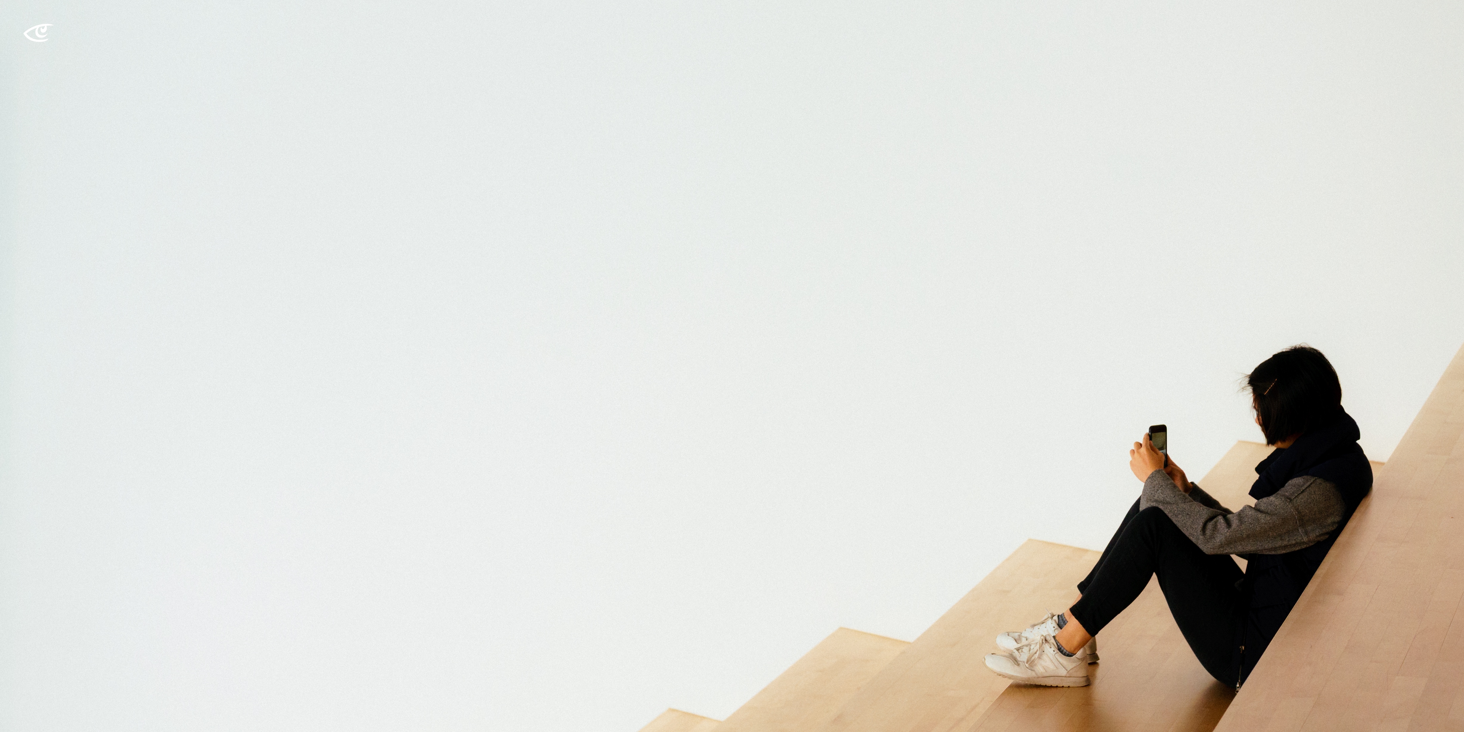Person sitting on light wooden stairs against a plain white wall, legs bent and holding a smartphone, with large empty space on the left side of the frame.
