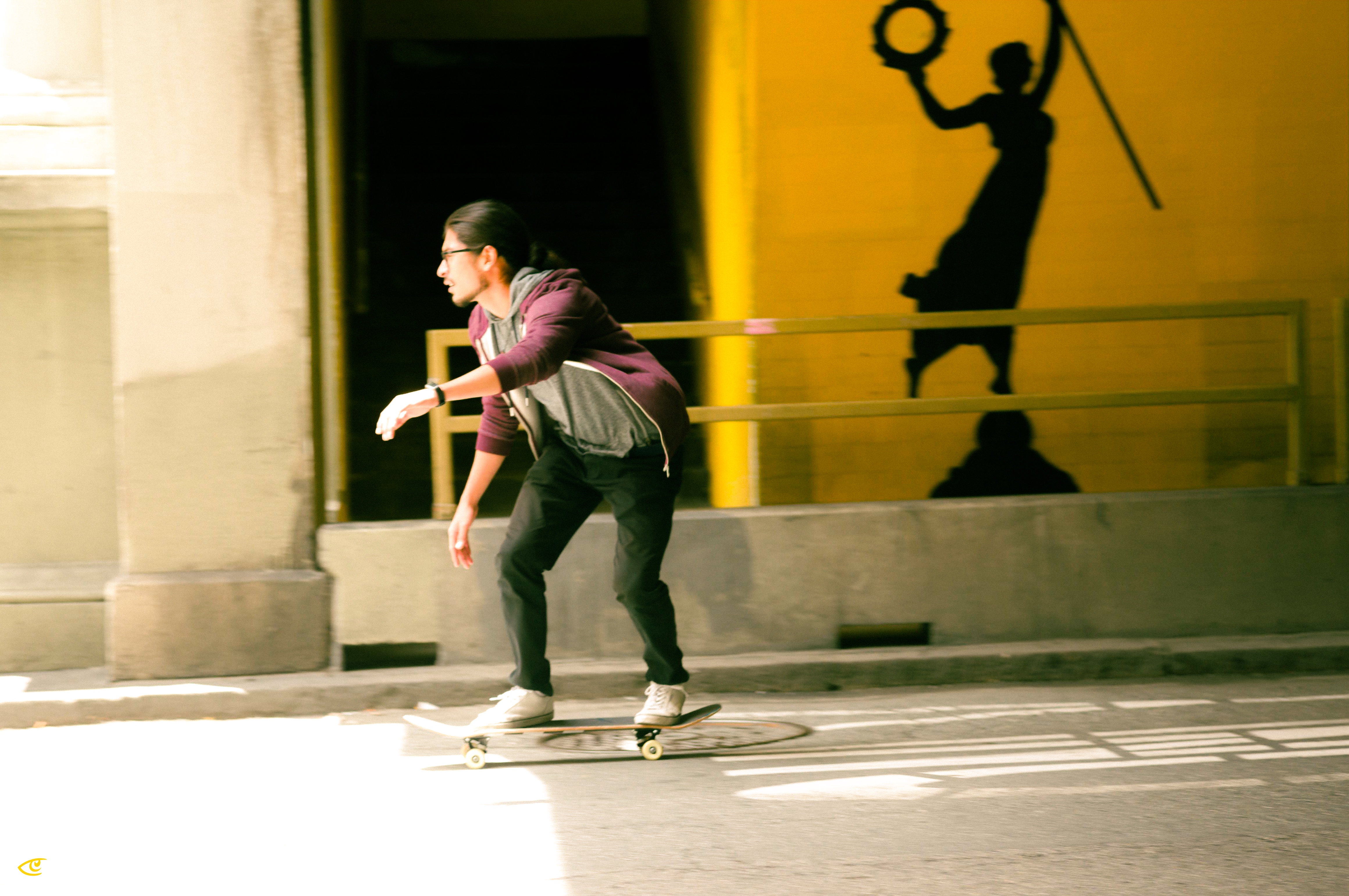 Person skateboarding on a sunlit street past a yellow wall with a railing, casting a shadow that resembles a figure holding a circular object and a stick.