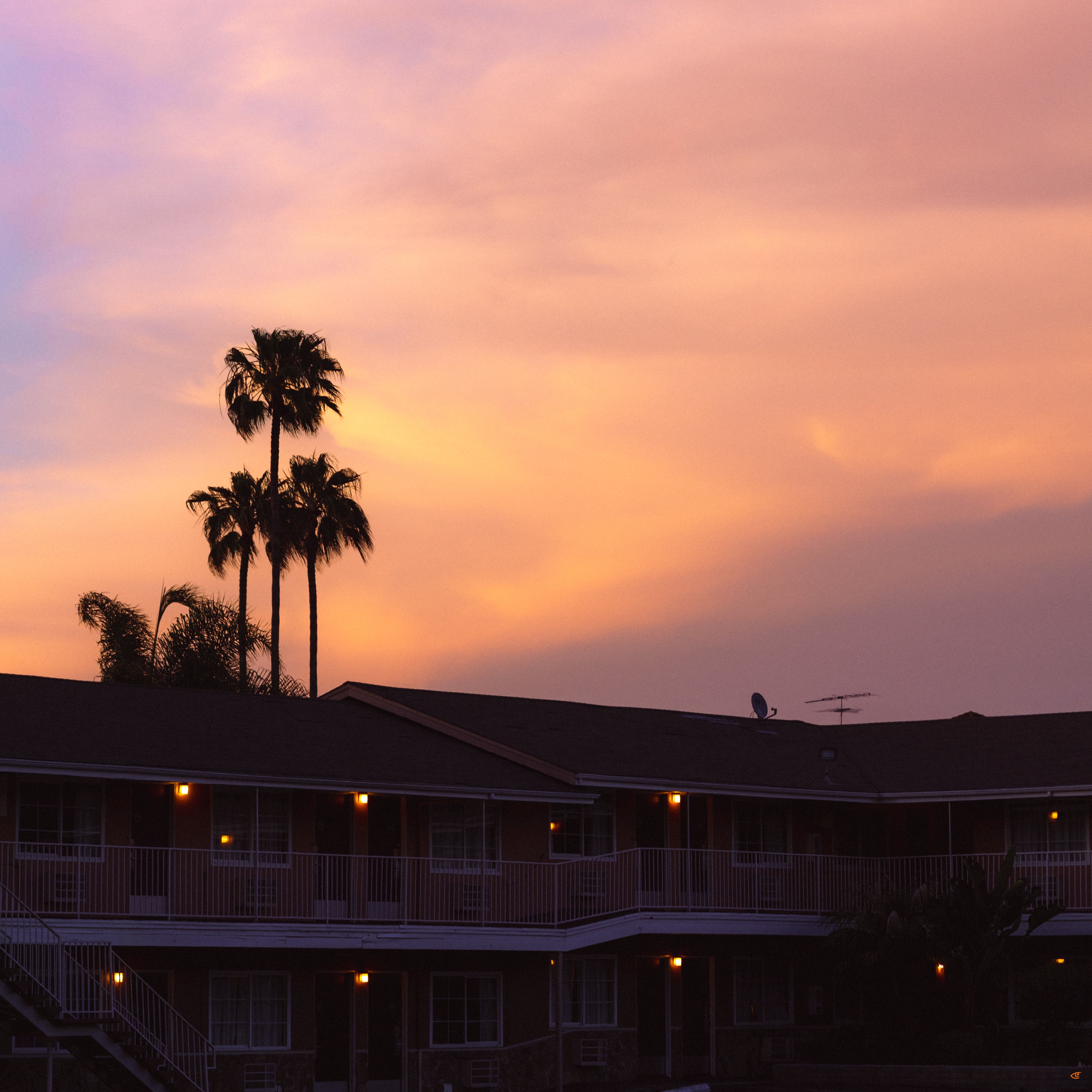 Silhouetted two-story motel with lit exterior hallway lights and railings, palm trees rising above the roofline, and a pink-and-orange sunset sky with scattered clouds.