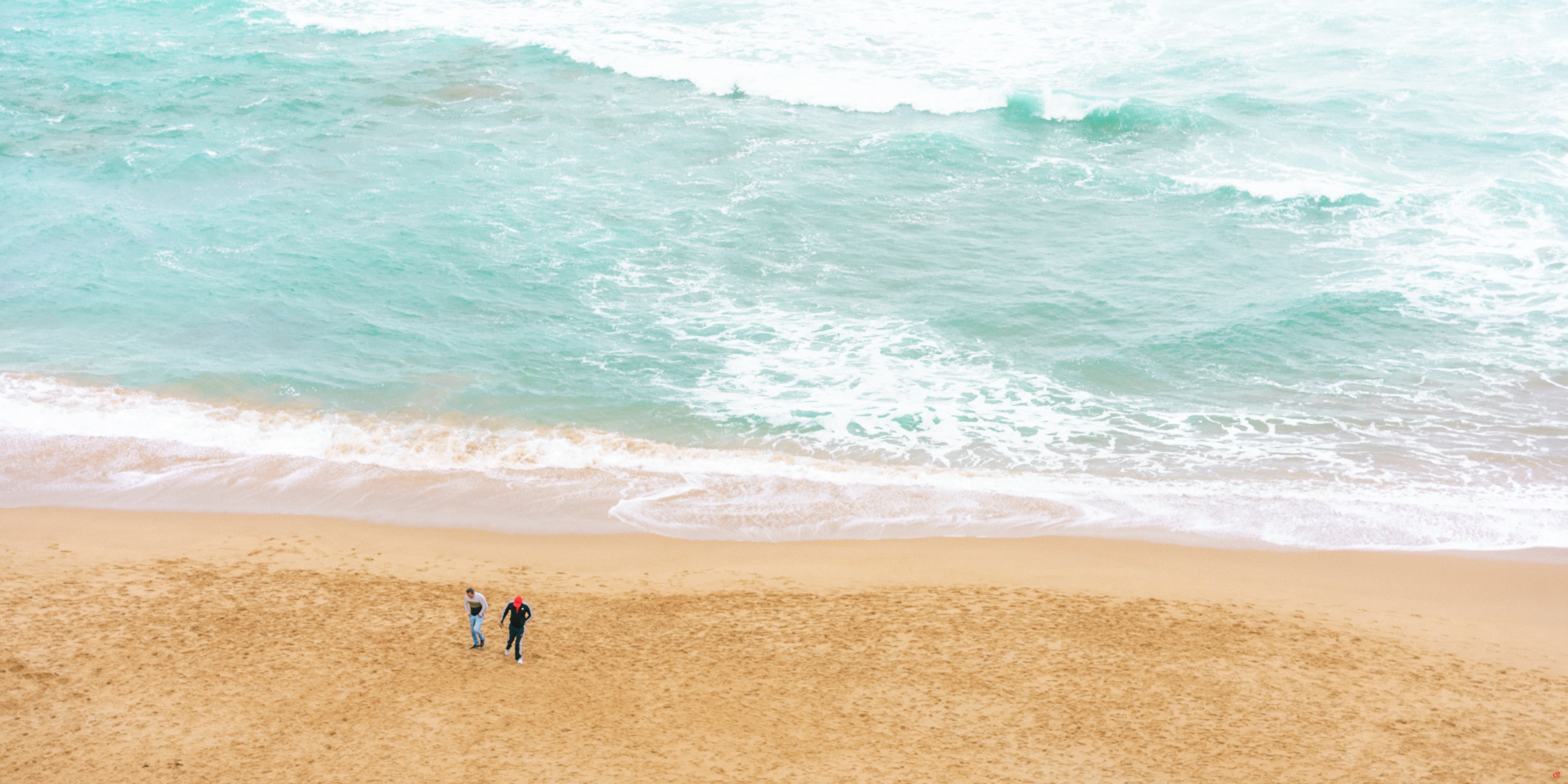 Two people standing on a wide sandy beach near the shoreline, with turquoise ocean waves and white foam filling the upper half of the image.