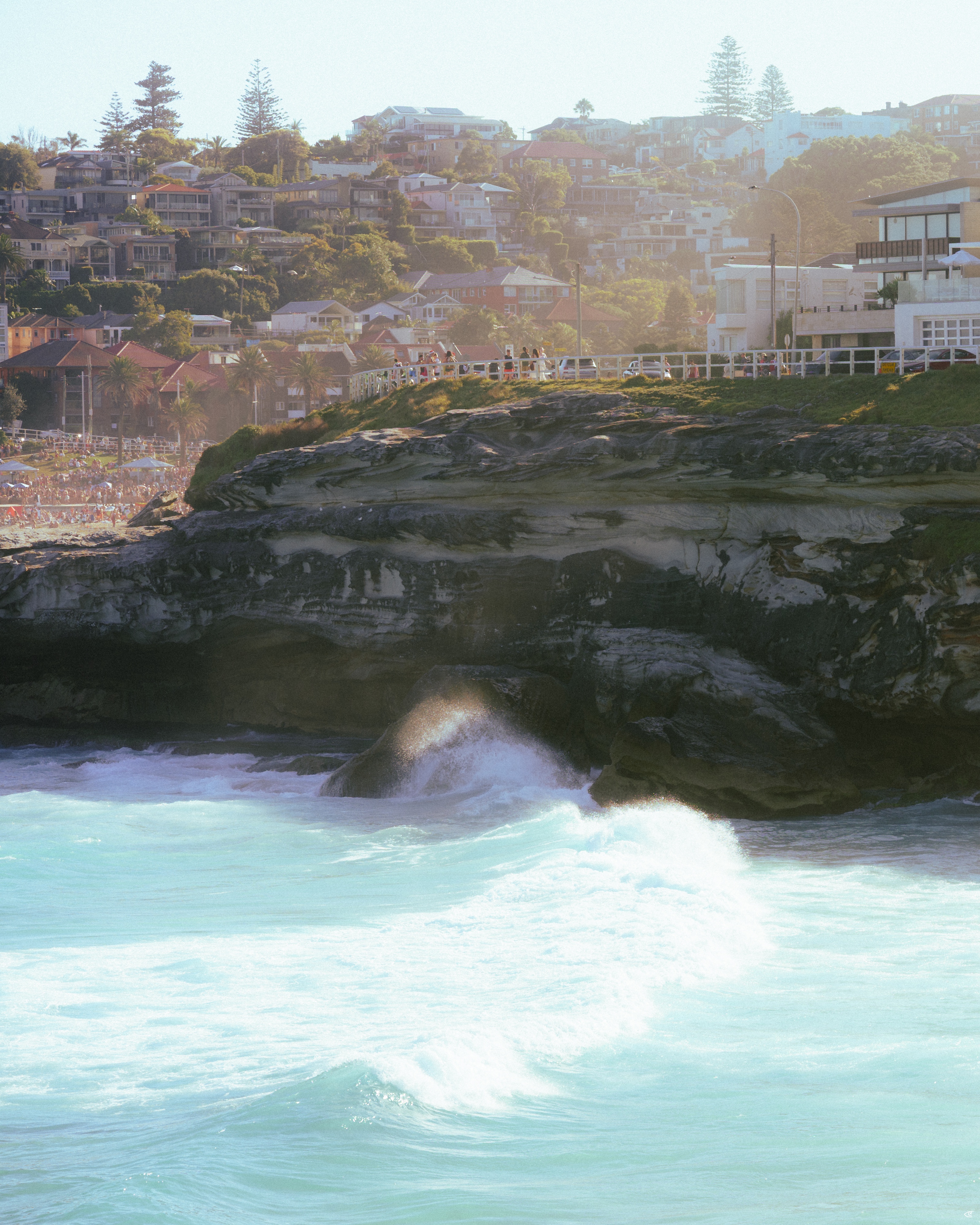 Waves crashing against a dark rocky cliff, with a seaside promenade and buildings along the edge, and a hillside neighborhood of houses and trees rising in the background.