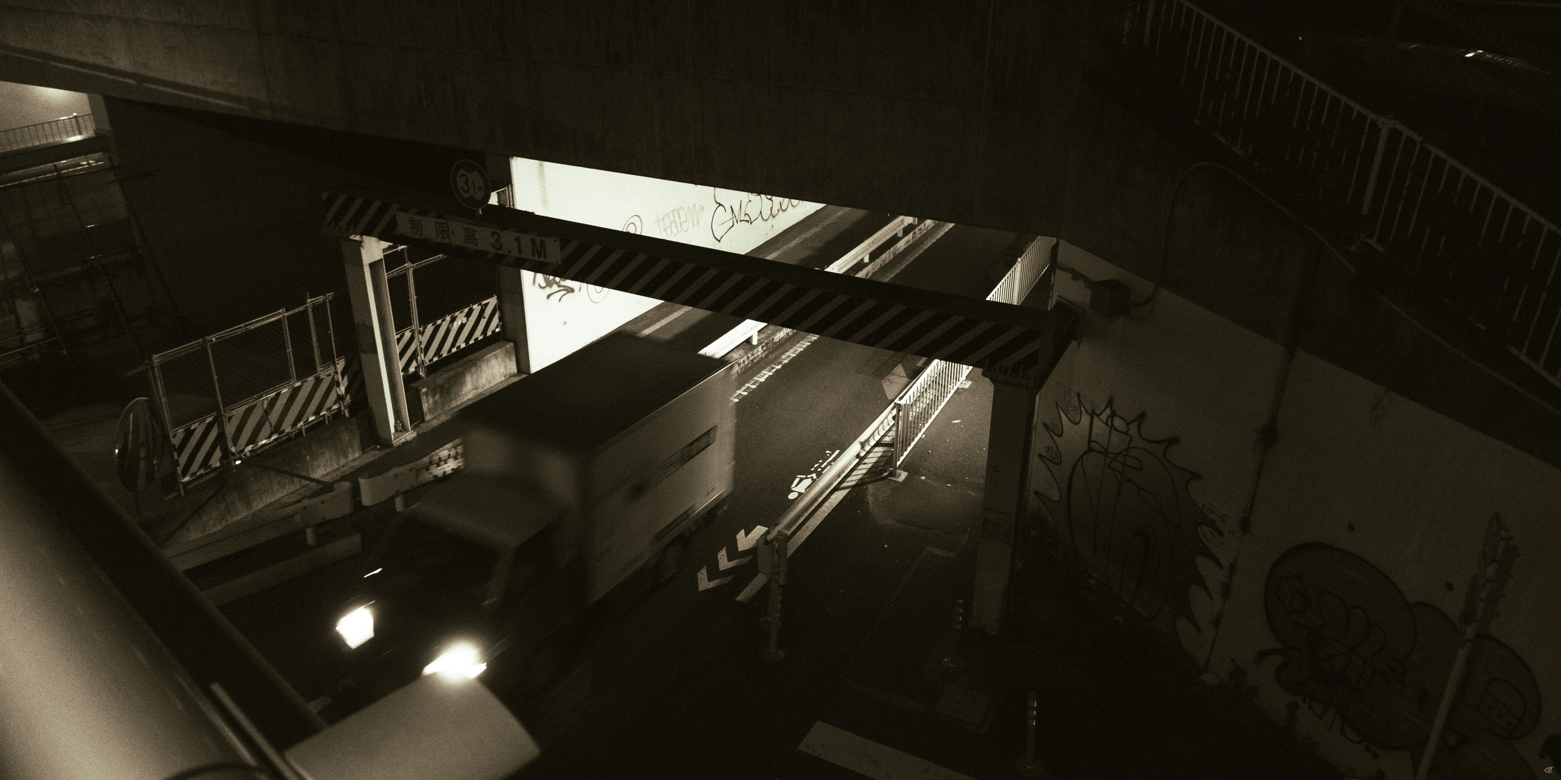 Overhead view of a dimly lit underpass with a truck passing beneath a metal beam structure, concrete walls covered in graffiti, and stairways and railings along both sides.