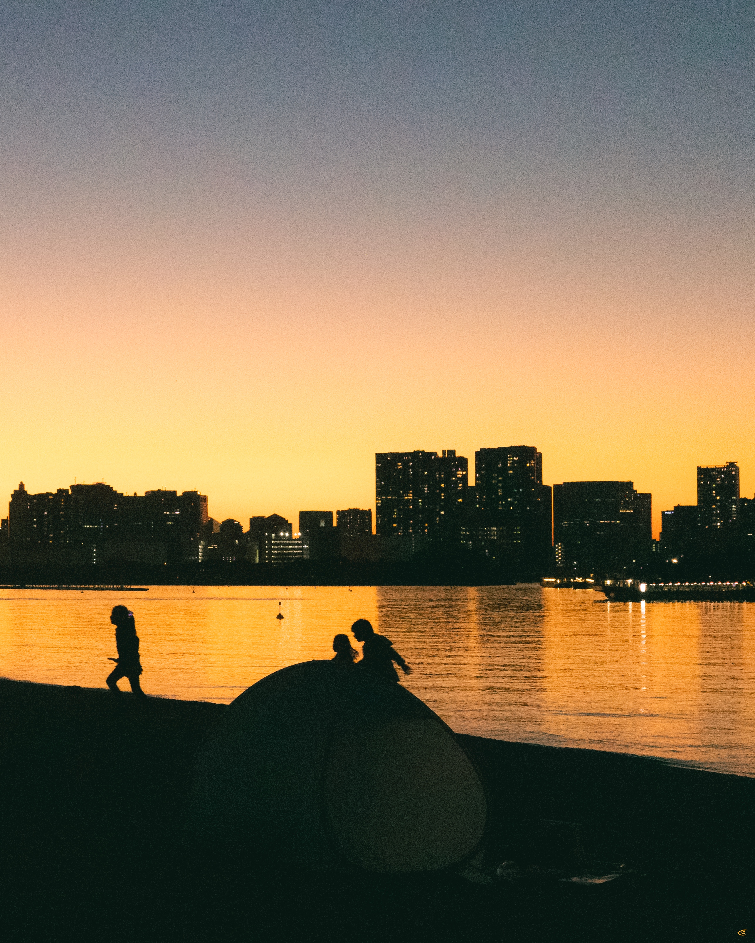 Silhouettes of three people on large rocks by a calm waterfront at sunset, with a city skyline and lit windows across the water under a yellow-to-blue gradient sky.