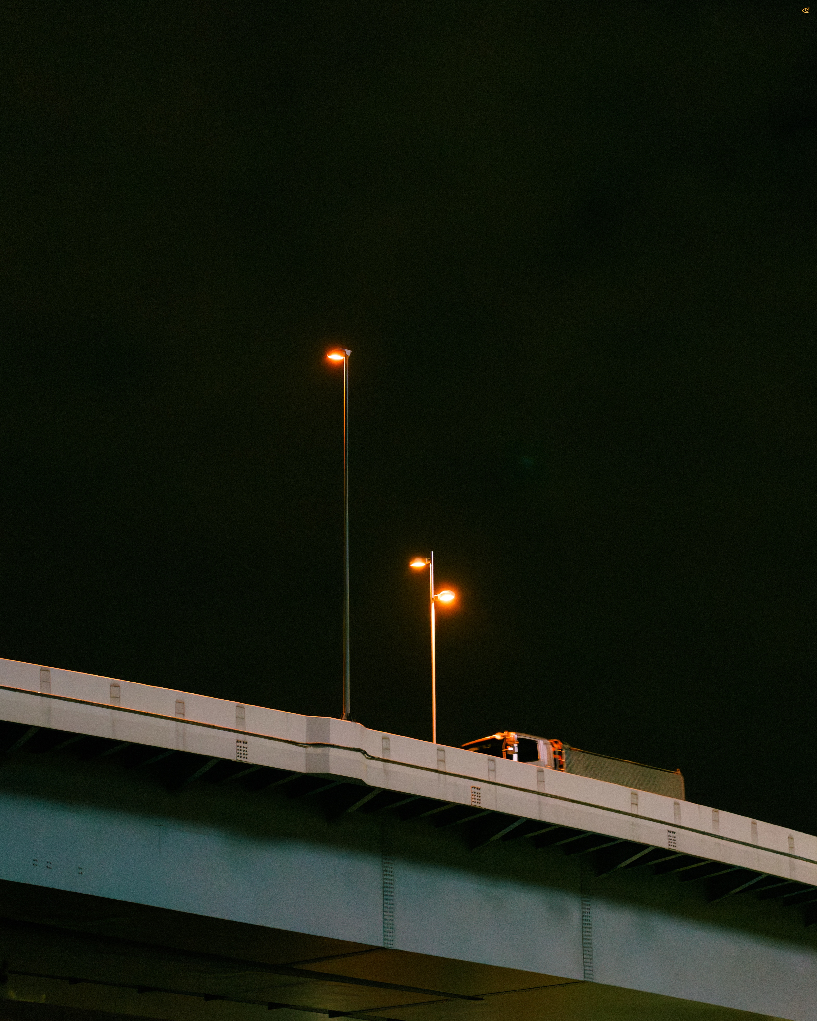 Elevated highway overpass at night with three tall streetlights glowing orange against a dark sky and a small orange maintenance vehicle parked near the right edge.