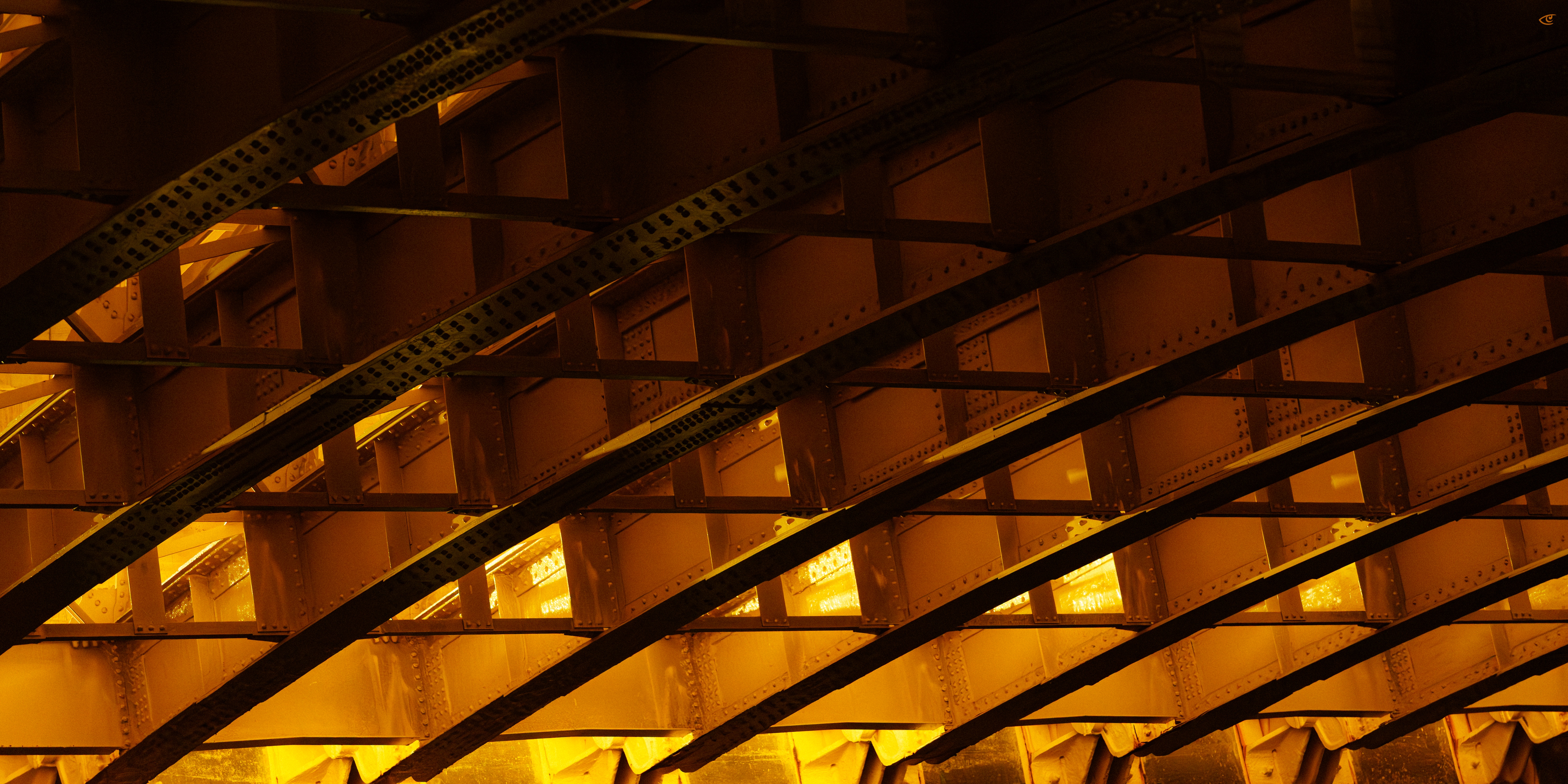 Underside of a steel bridge with repeating riveted beams and cross-bracing, intersected by several diagonal girders, lit by warm yellow-orange lights.