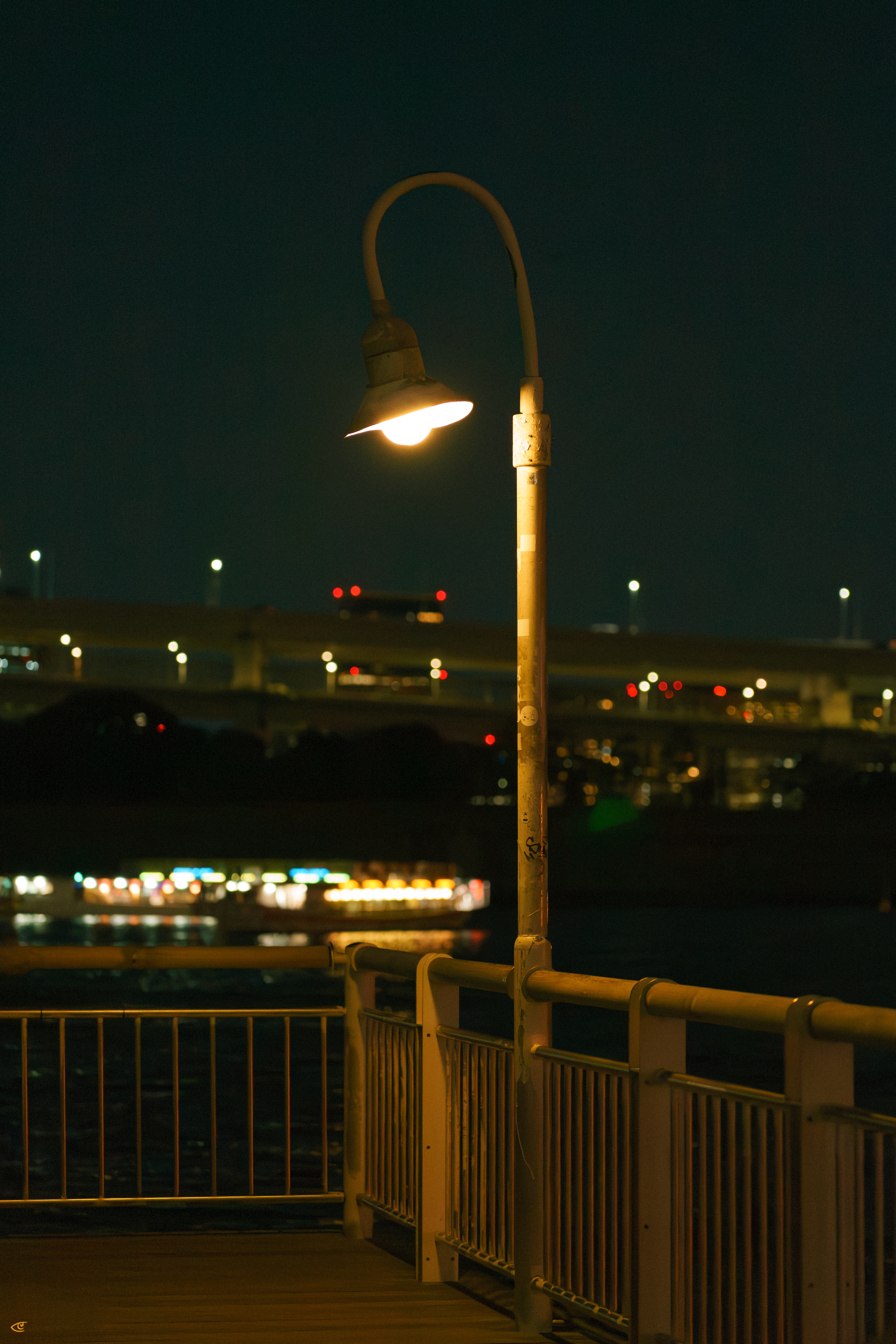 Curved streetlamp glowing above a metal-railed walkway at night, with blurred city lights and an elevated roadway in the background.