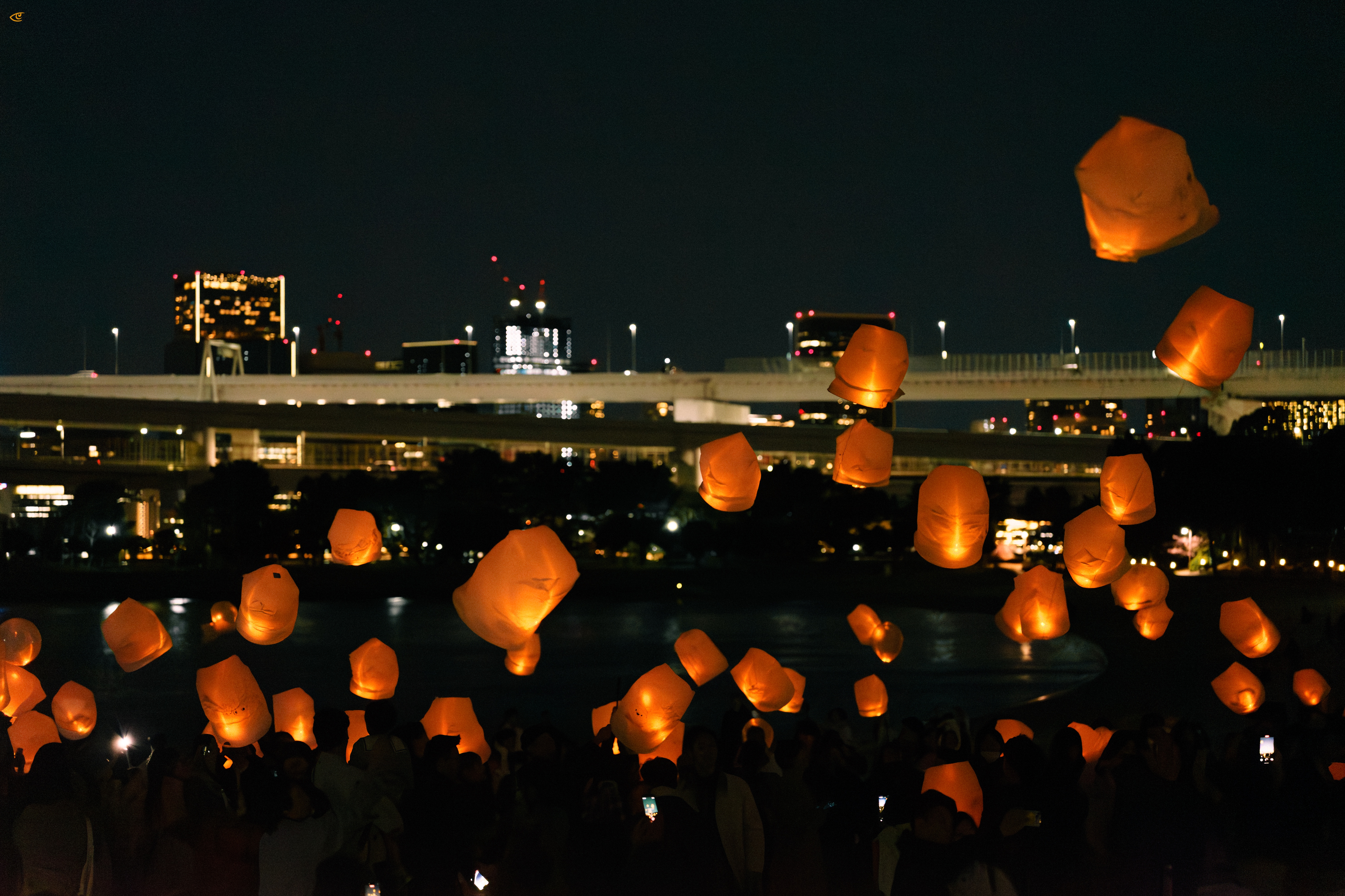 Orange paper sky lanterns floating above a crowd at night beside a river, with silhouetted people holding lanterns in the foreground and a lit bridge and city buildings in the background.