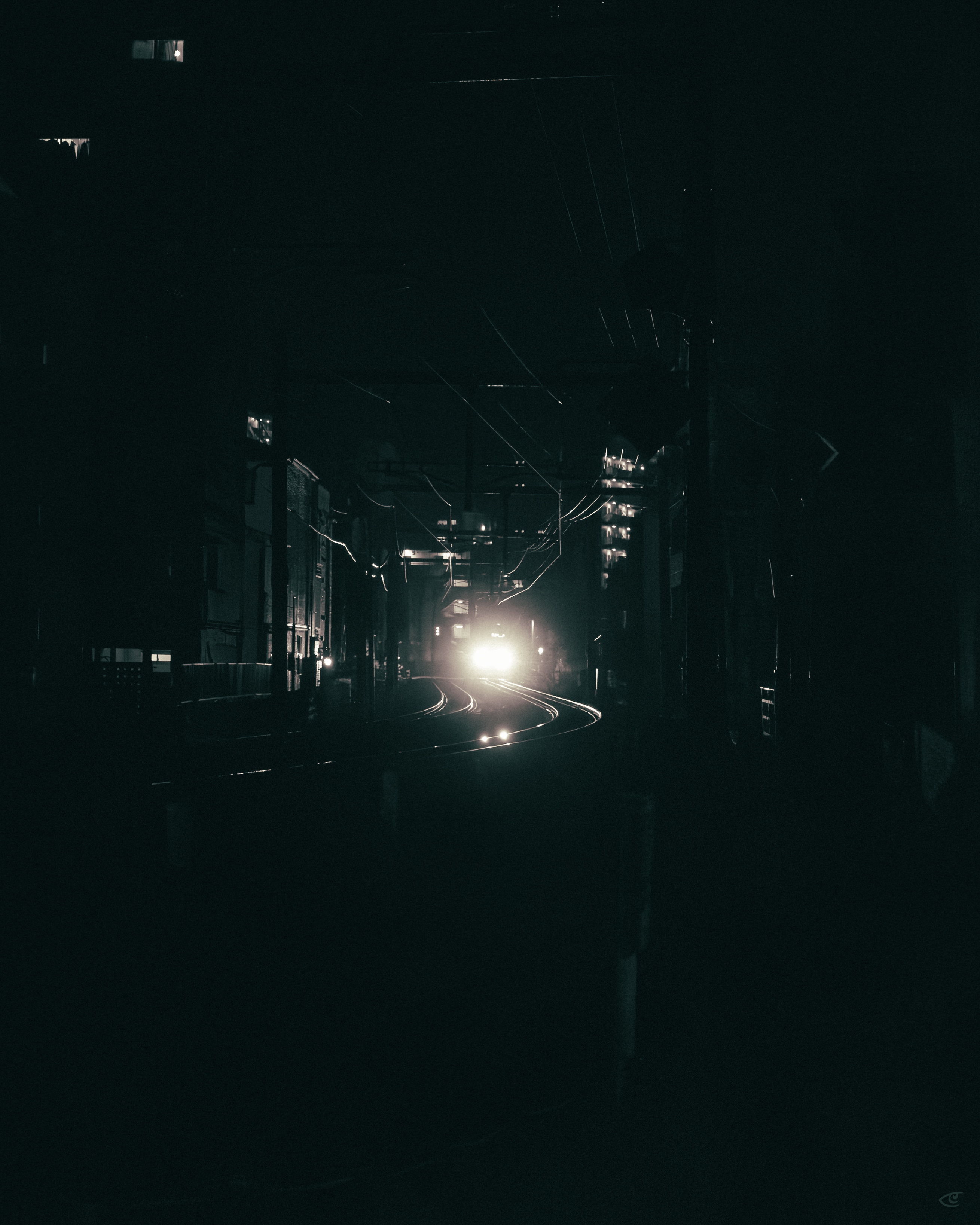 Dark nighttime view down a narrow urban rail corridor, with curving train tracks leading toward bright headlights in the distance, buildings and utility wires lining both sides, and light rain streaks visible.