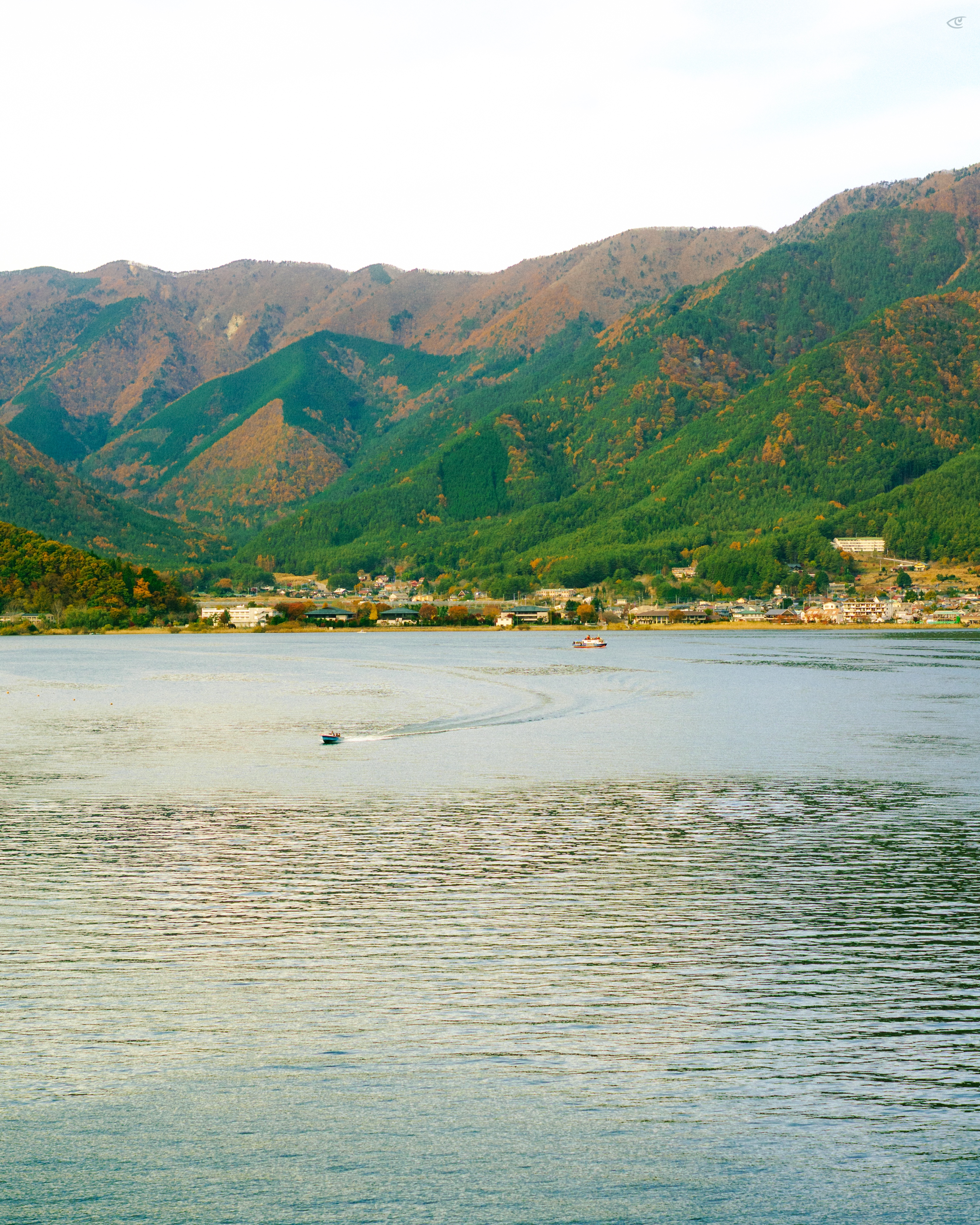 Wide view across a lake with rippled water and two small boats leaving curved wakes, a shoreline town in the distance, and forested mountains rising behind under a pale sky.