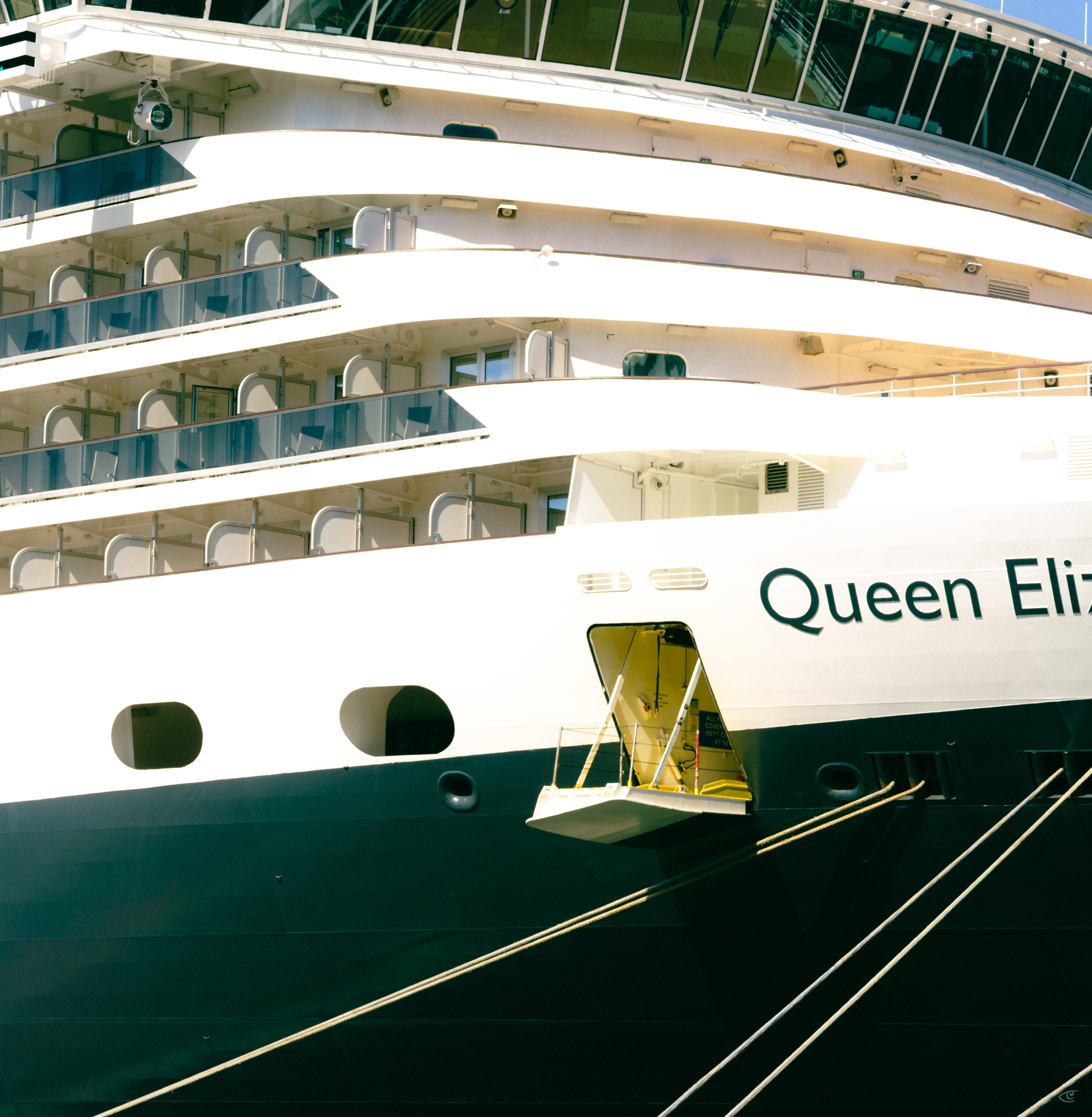 Close-up of a cruise ship’s bow showing stacked balcony decks above a black hull, an open side door with a small platform, thick mooring lines stretching diagonally, and the partial name “Queen Eli” painted on the white side.