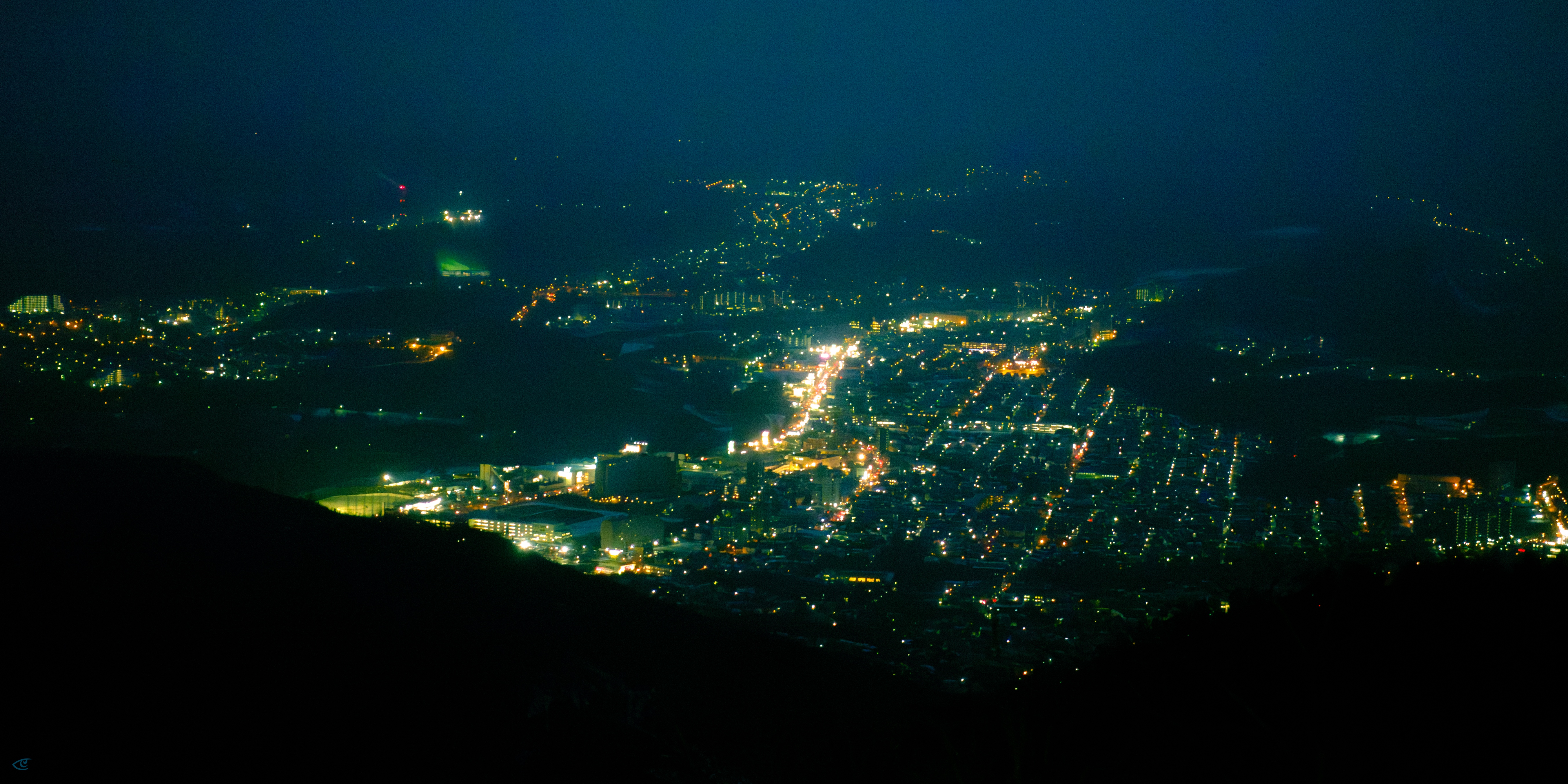 Aerial nighttime view of a city with streetlights forming a grid and a bright main road running through the center, surrounded by dark hills in the foreground and a dark sky above.