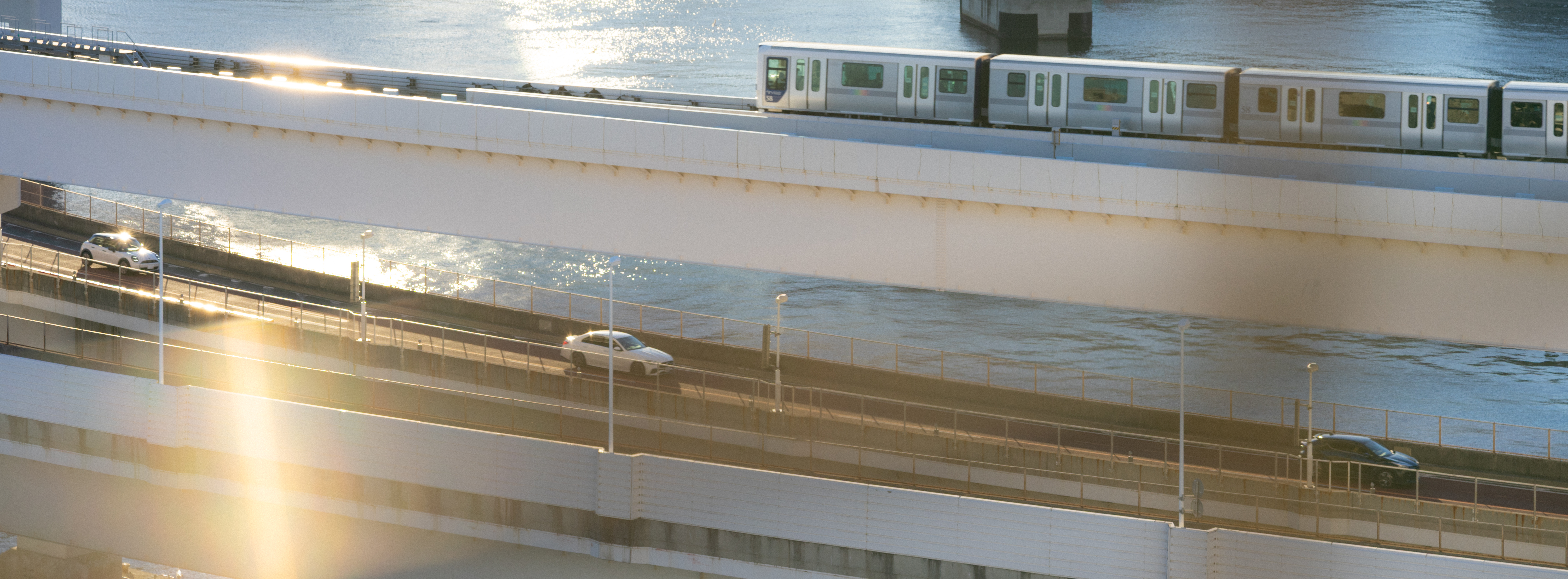 Elevated train traveling on a bridge above a river. Below the train, there is a roadway with two visible cars, separated from the train bridge by another structure. The sun creates a lens flare, casting a warm glow on the scene.