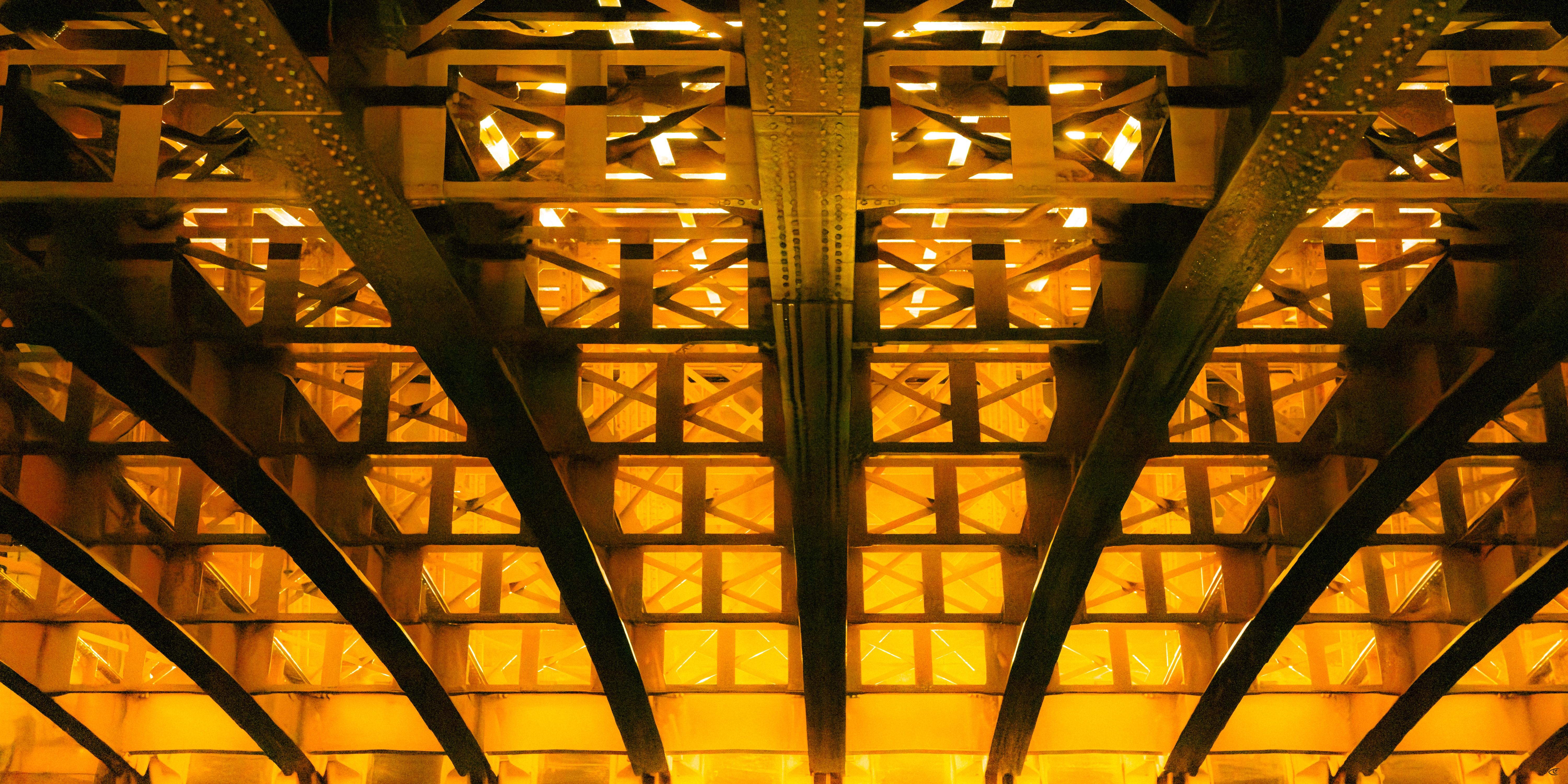 Upward view of a steel bridge underside with intersecting girders and riveted beams, lit in warm yellow-orange light with symmetrical repeating cross-bracing.