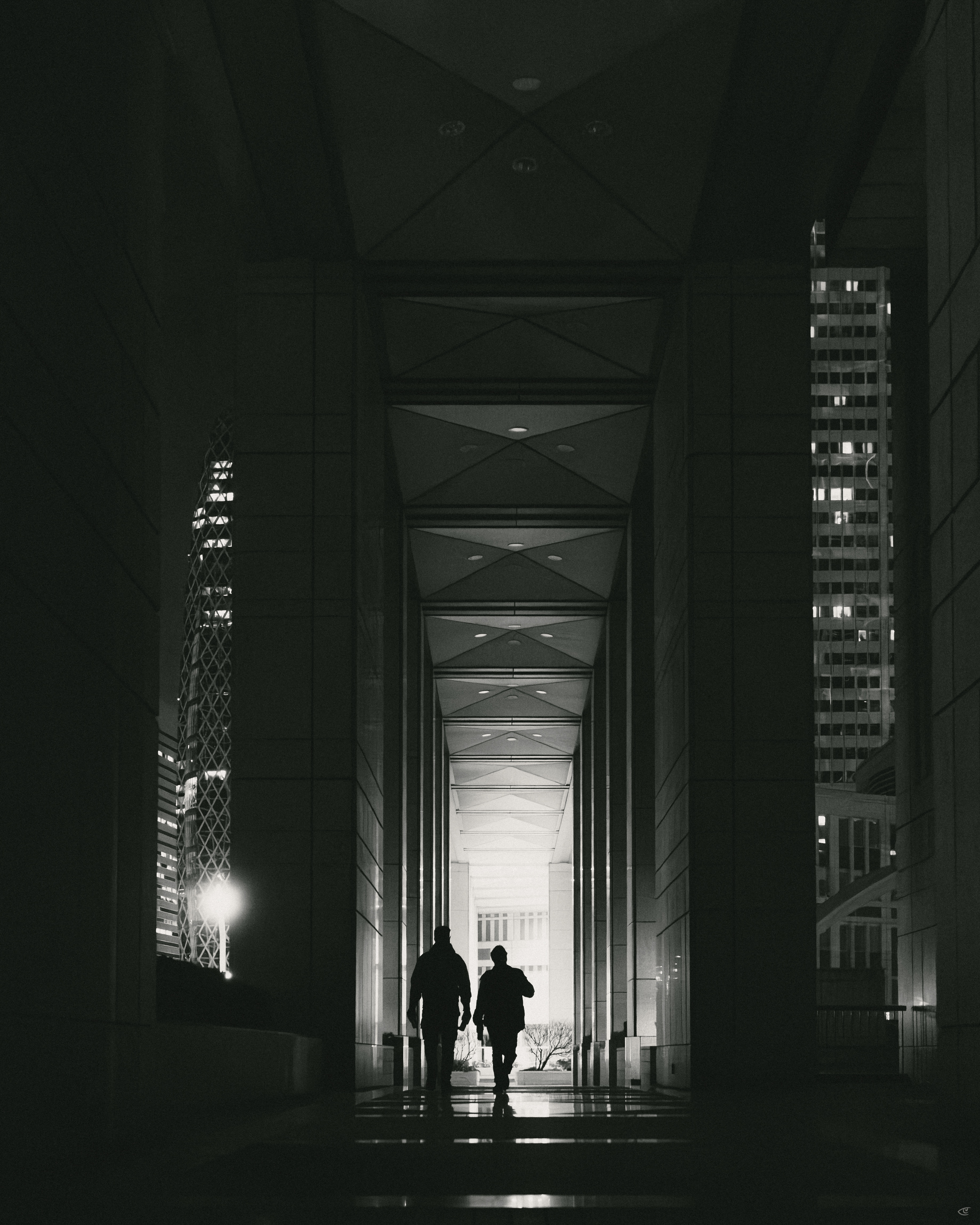 Two silhouetted pedestrians walking through a tall, covered urban passage with repeating ceiling panels and columns, lit from a bright opening at the far end; high-rise buildings with lit windows visible on both sides at night.