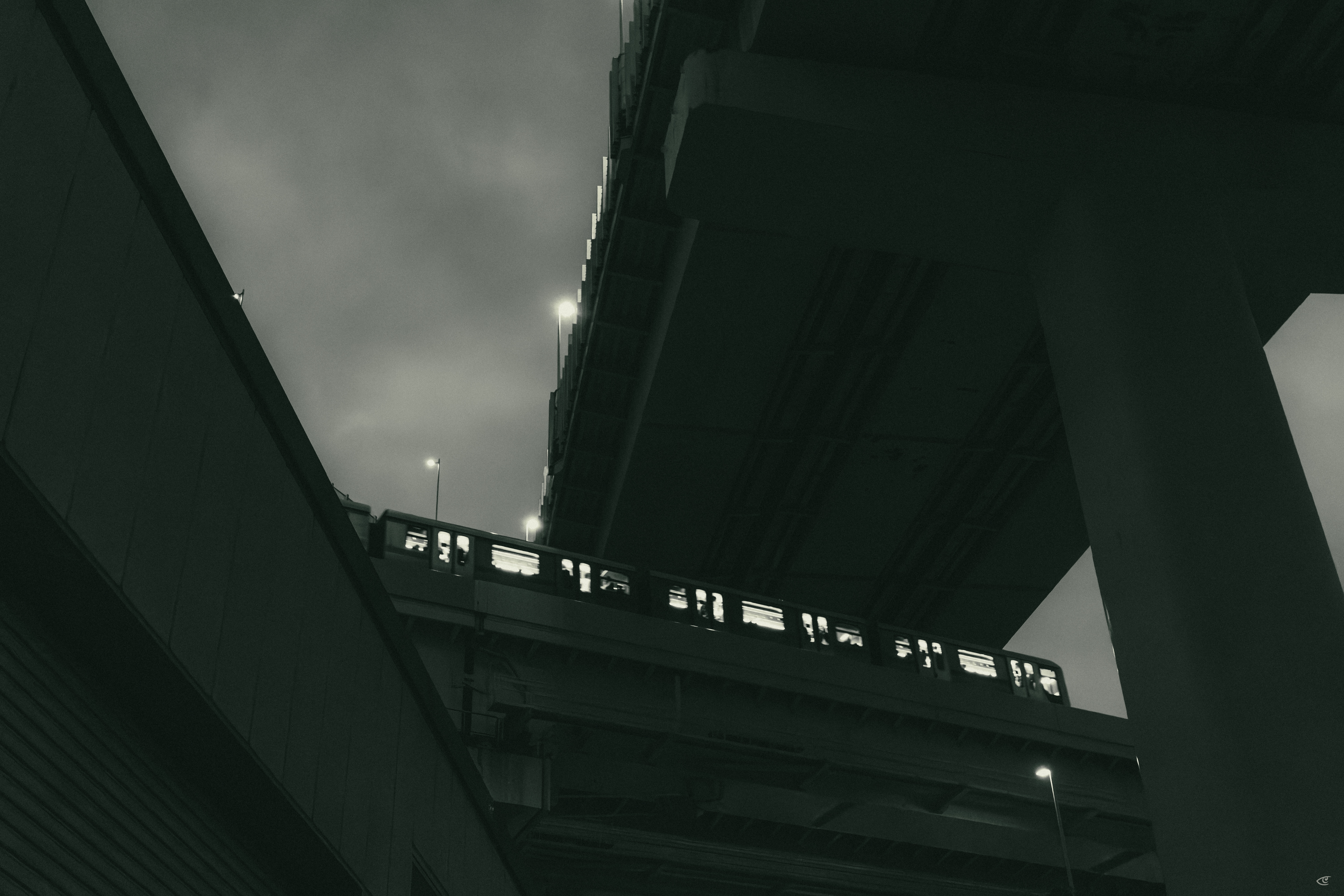 Low-angle view between two elevated overpasses, with a commuter train crossing a bridge section above; tall support columns frame the right side under an overcast sky.