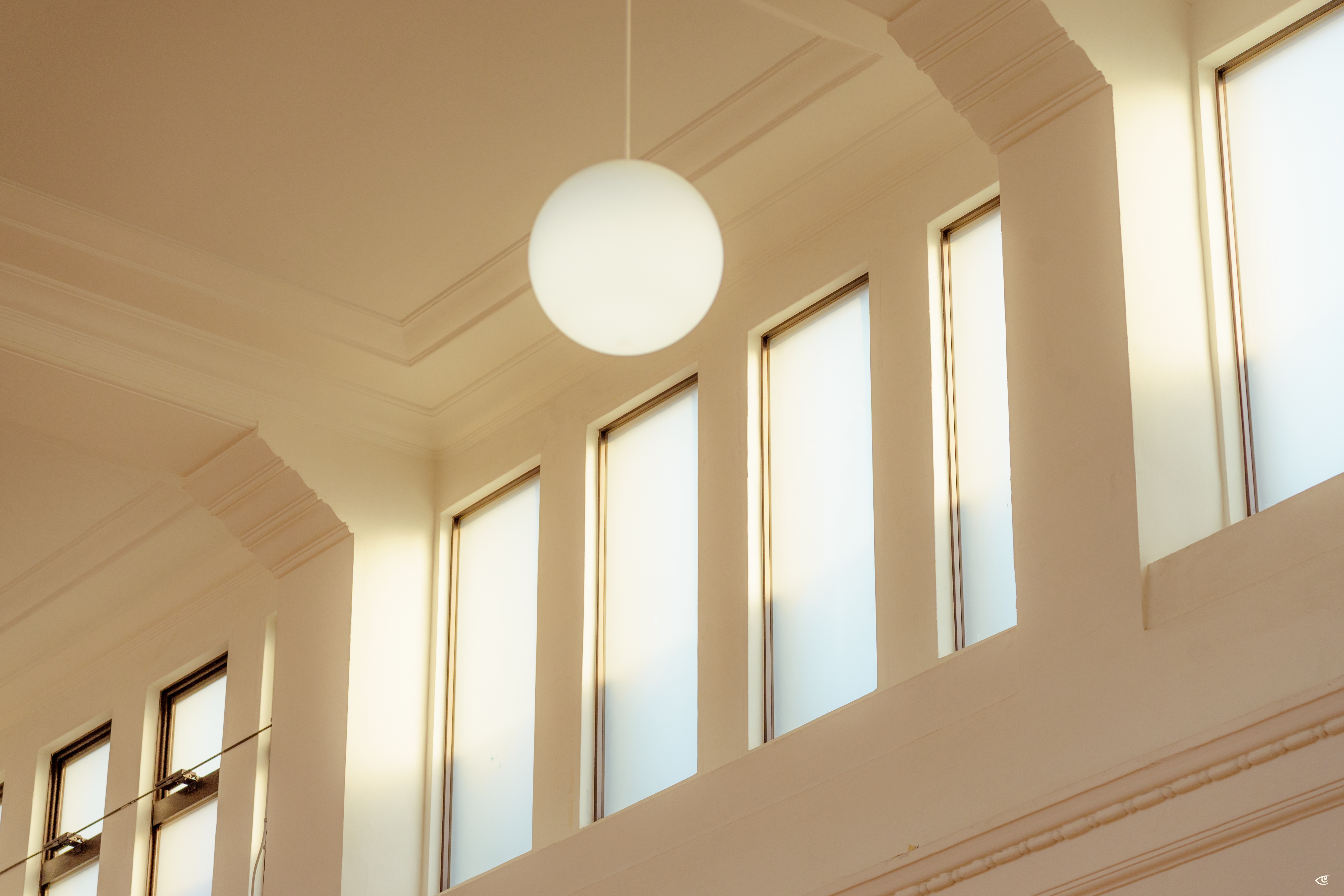 Round white pendant light hanging from a high ceiling above a row of tall, narrow rectangular windows set in a cream-colored wall with decorative molding.