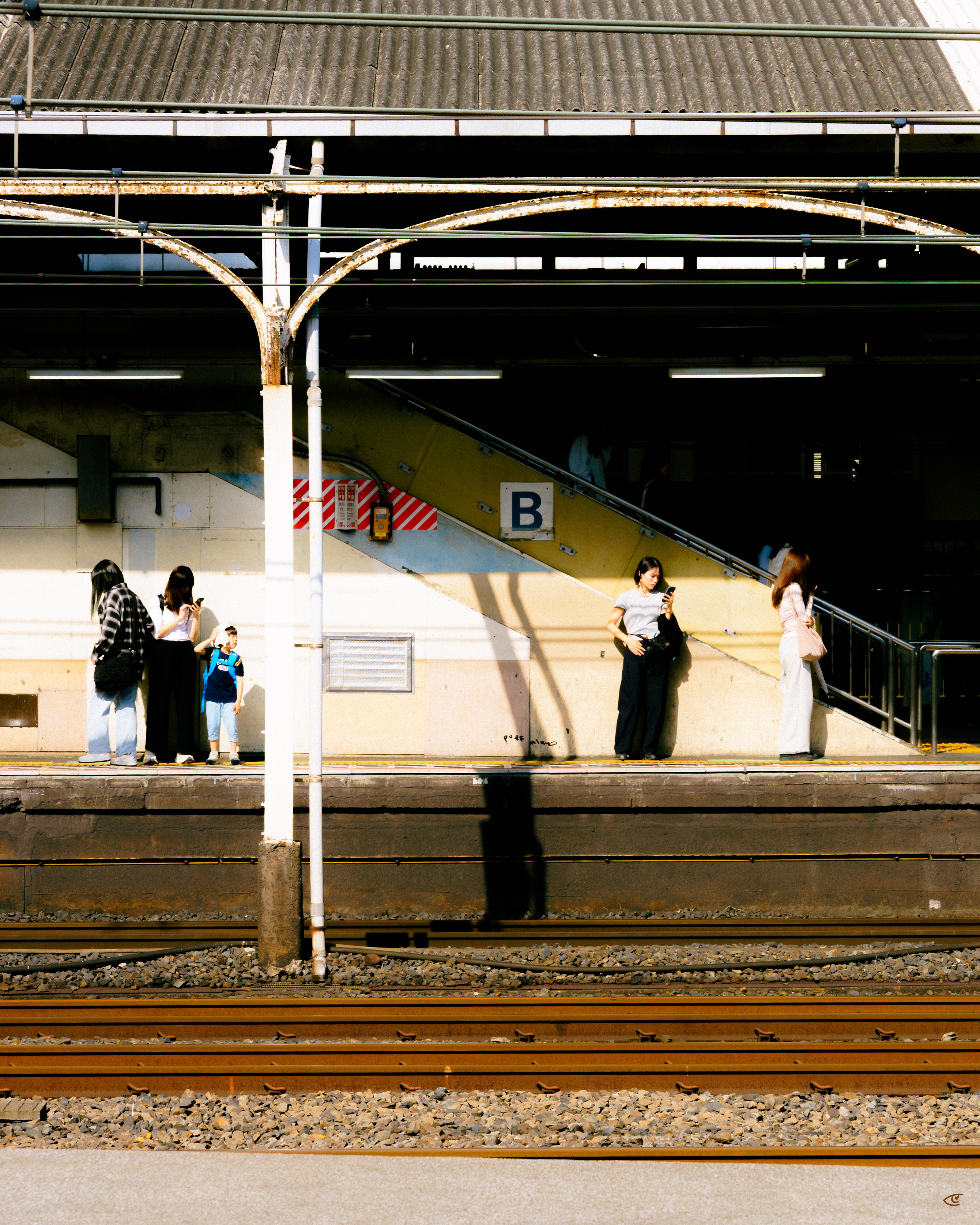 Train station platform with several people waiting, a child in a blue outfit, and two women looking at phones; overhead metal canopy and columns, a stairway marked with a large “B,” and train tracks in the foreground with long shadows cast across the platform.