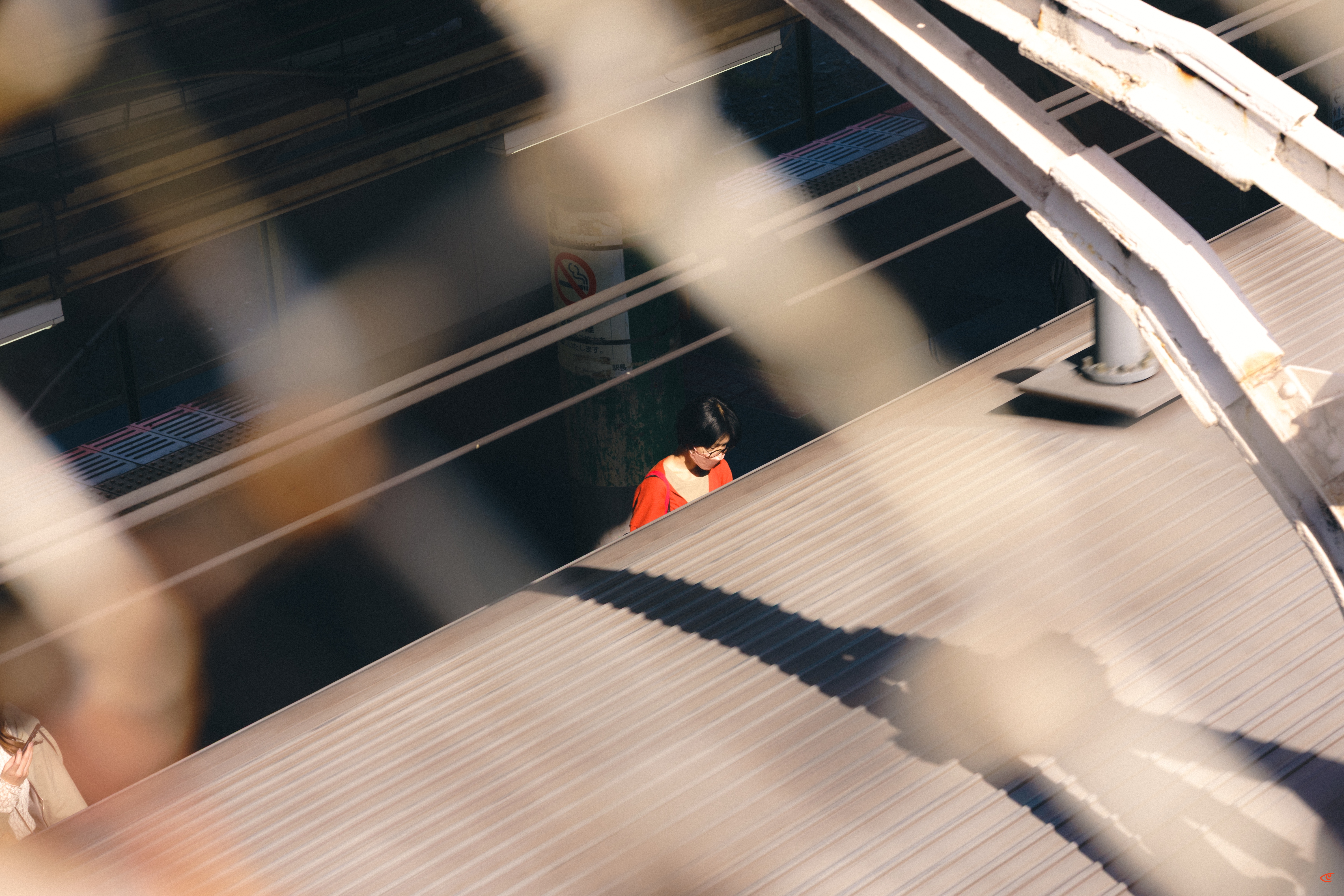 Overhead view through a blurred chain-link fence of a person in a red top walking along a narrow passage beside a large corrugated metal roof and white structural beams.