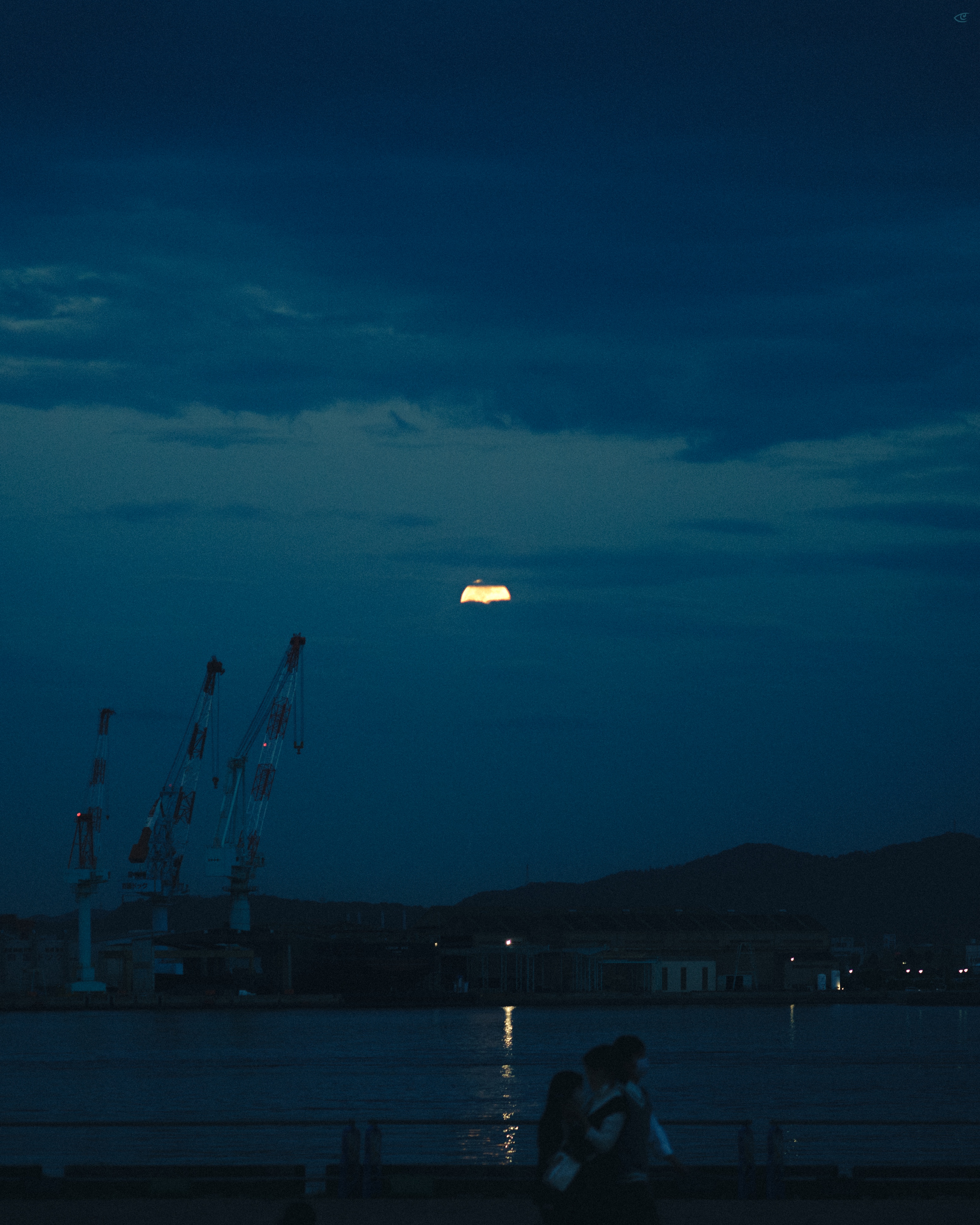 Dark, cloud-filled sky over a waterfront at dusk, with a thin sliver of sun near the horizon reflecting on the water; cranes and industrial buildings on the left, low hills on the right, and two blurred people silhouetted in the foreground near the shore.
