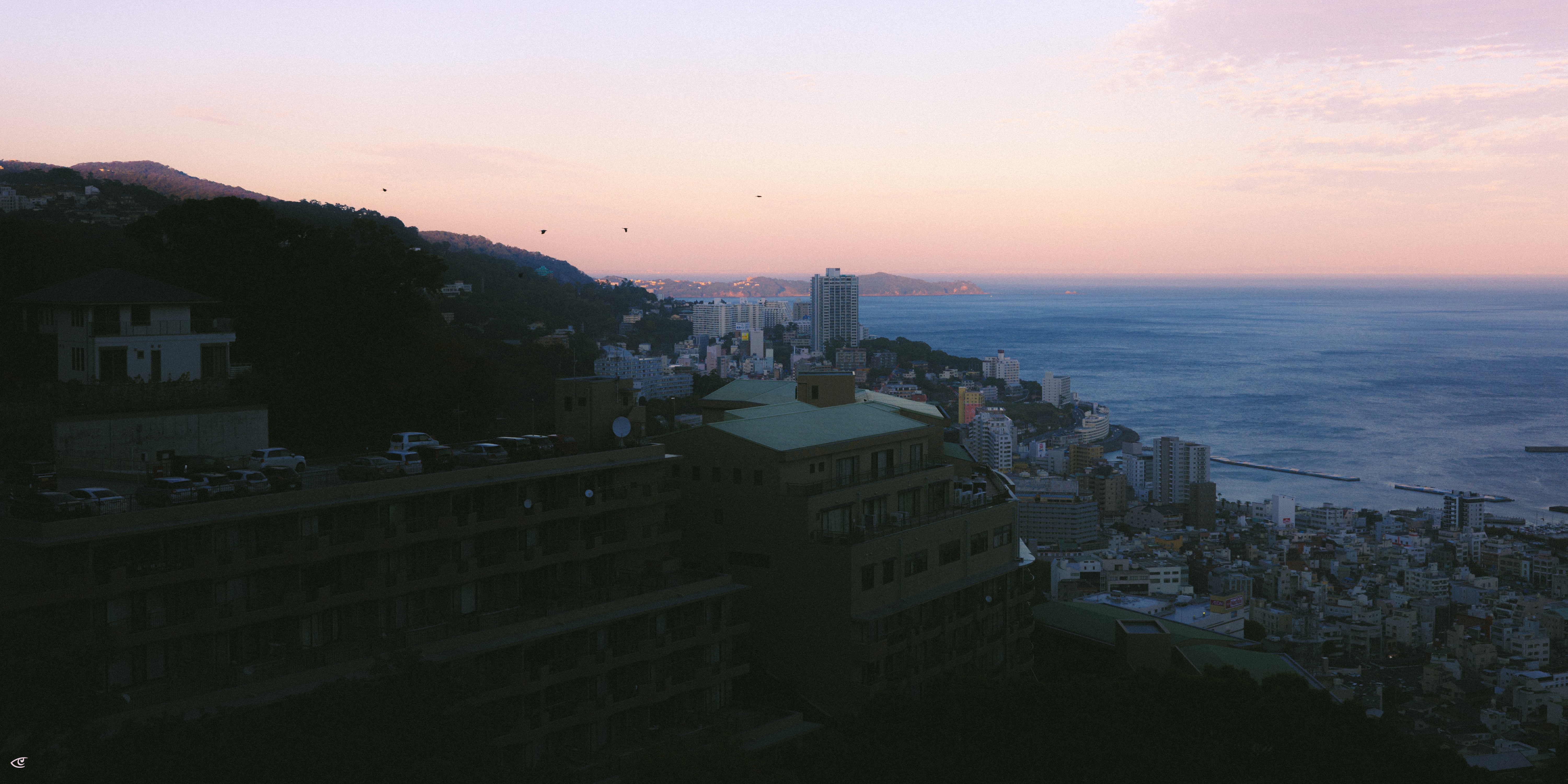 Coastal cityscape at dusk viewed from a hillside, with a large dark building in the foreground, densely packed buildings along the shoreline, and the ocean stretching to the horizon under a pink and blue sky.