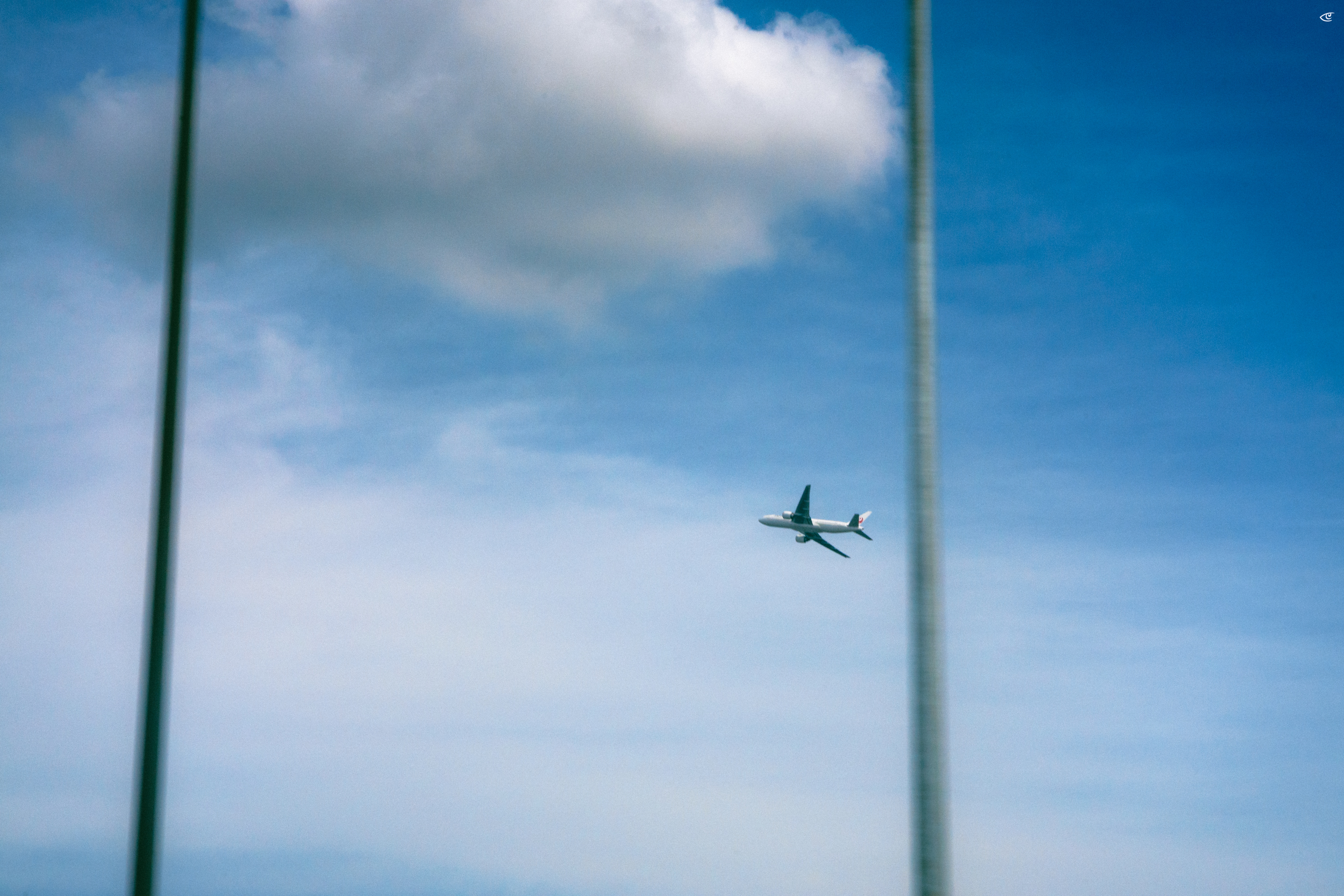 Small airplane flying across a blue sky with a large white cloud near the top left, framed by two tall out-of-focus vertical poles on the left and right edges.
