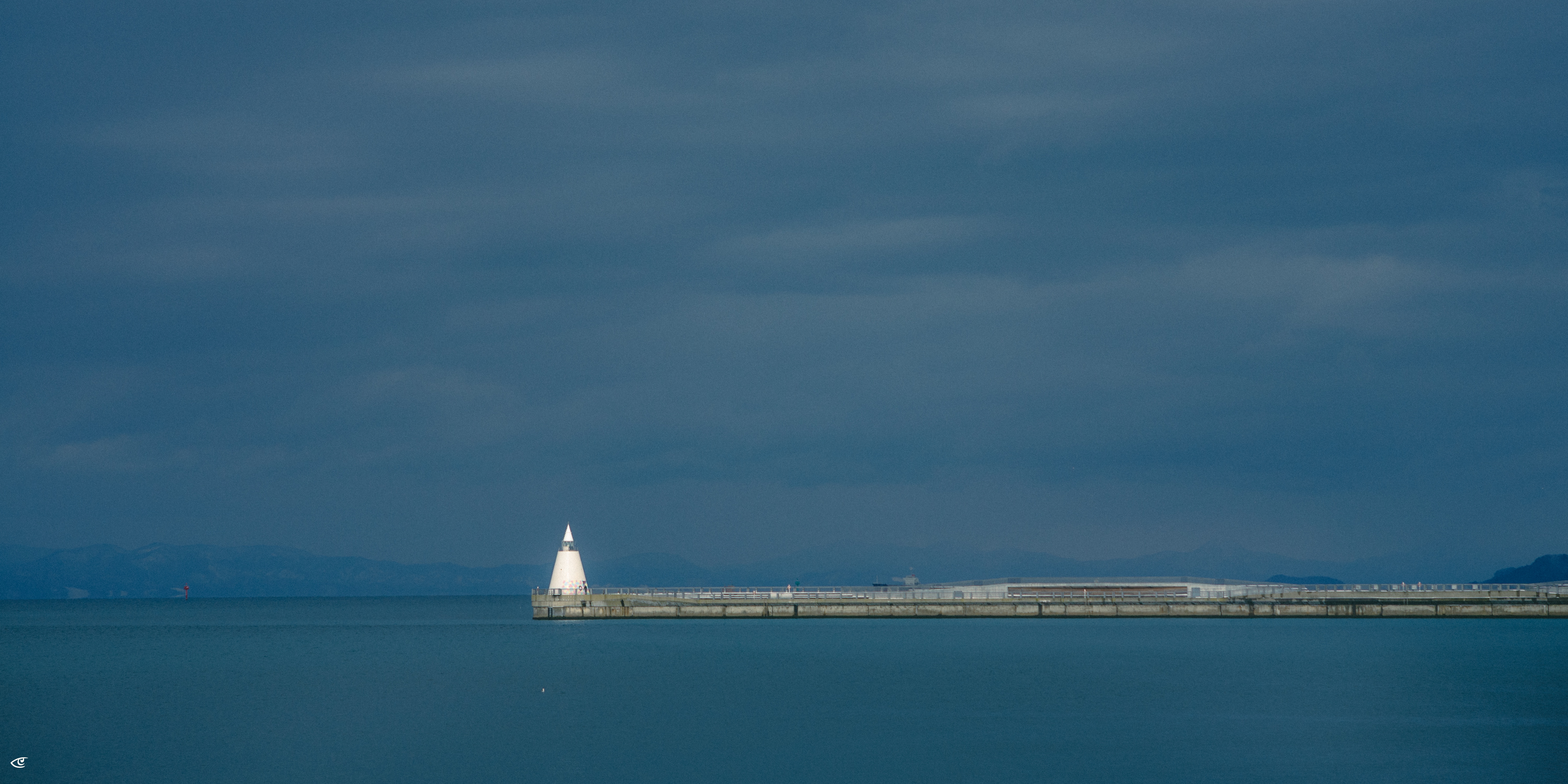 White lighthouse at the end of a long concrete breakwater extending from right to left across calm blue water, with dark cloudy sky and faint distant mountains on the horizon.