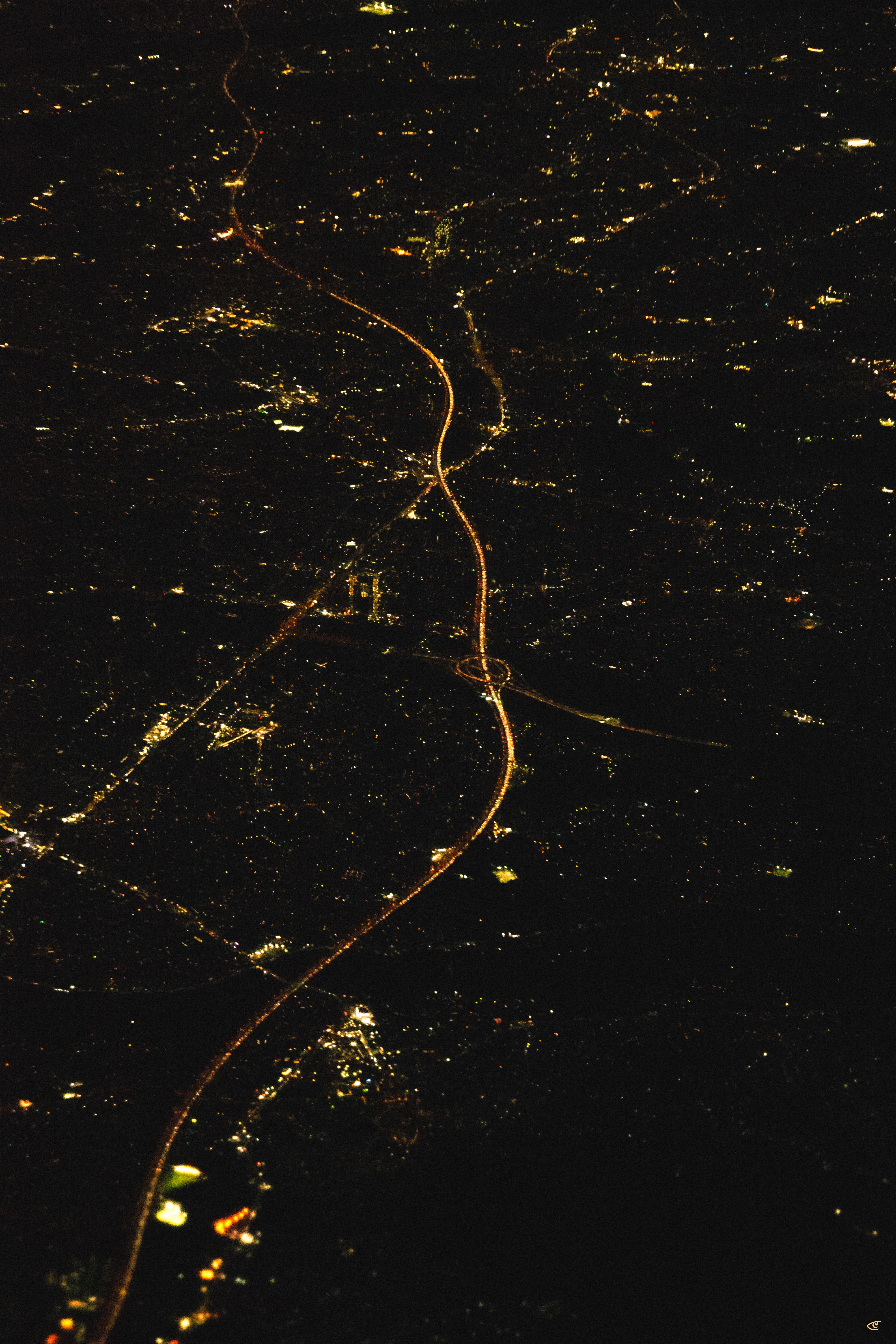 Aerial nighttime view of a city with scattered clusters of yellow lights; a brightly lit highway runs vertically through the center, curving and crossing an interchange near the lower middle, with dark areas and faint cloud shadows surrounding the illuminated streets.