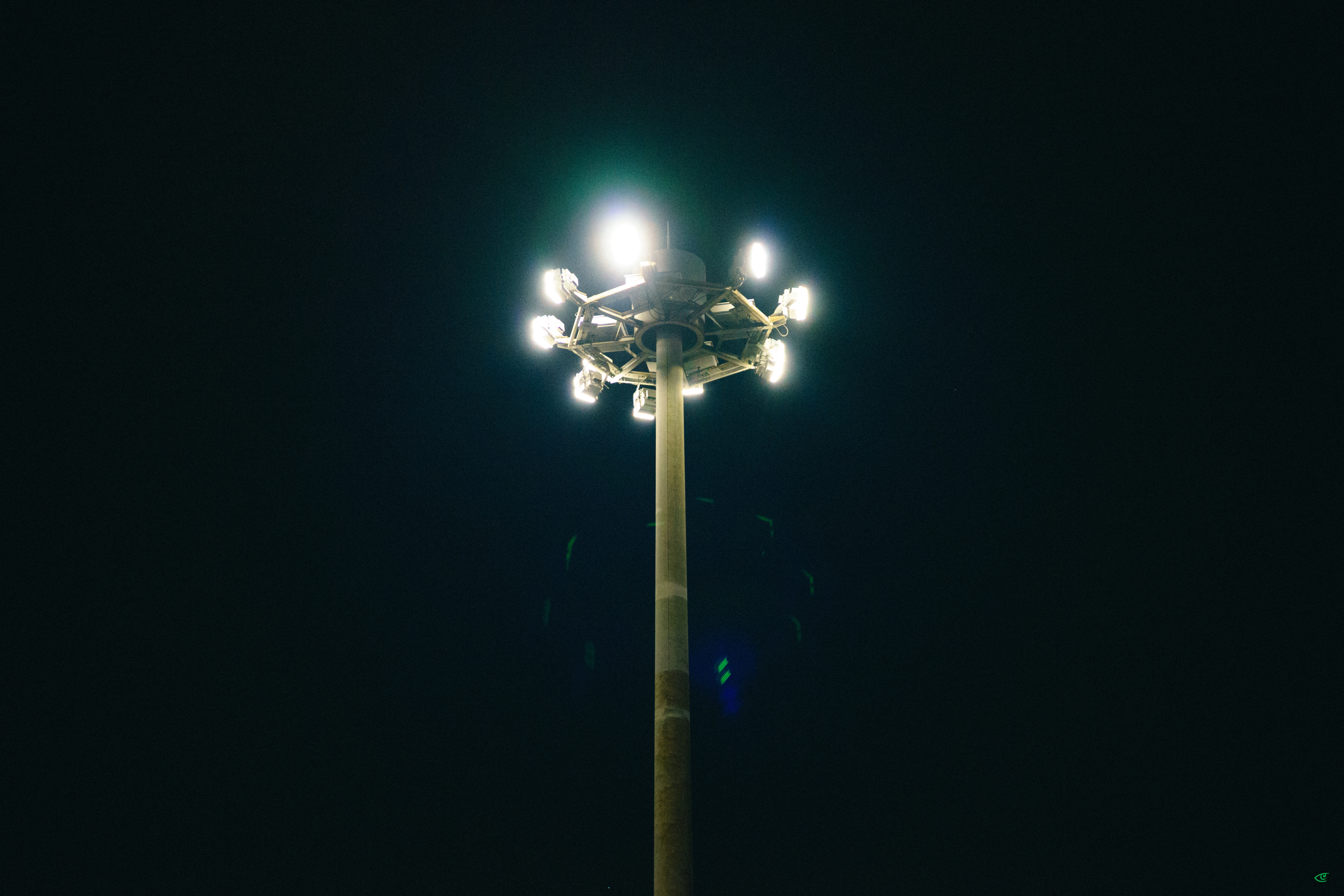 Tall stadium light pole centered against a dark night sky, with multiple bright floodlights at the top creating glare and lens flare.