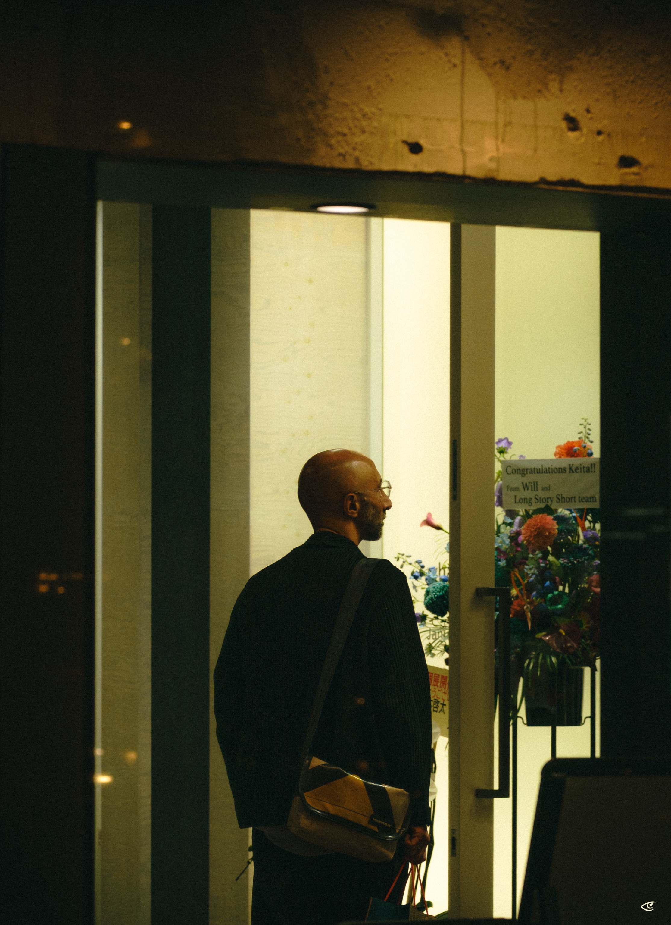Man standing in profile outside a glass doorway at night, wearing glasses and a dark coat with a shoulder bag, holding shopping bags; inside the brightly lit entryway are tall flower arrangements and a sign reading “Congratulations Keita!! … Long Story Short team.”