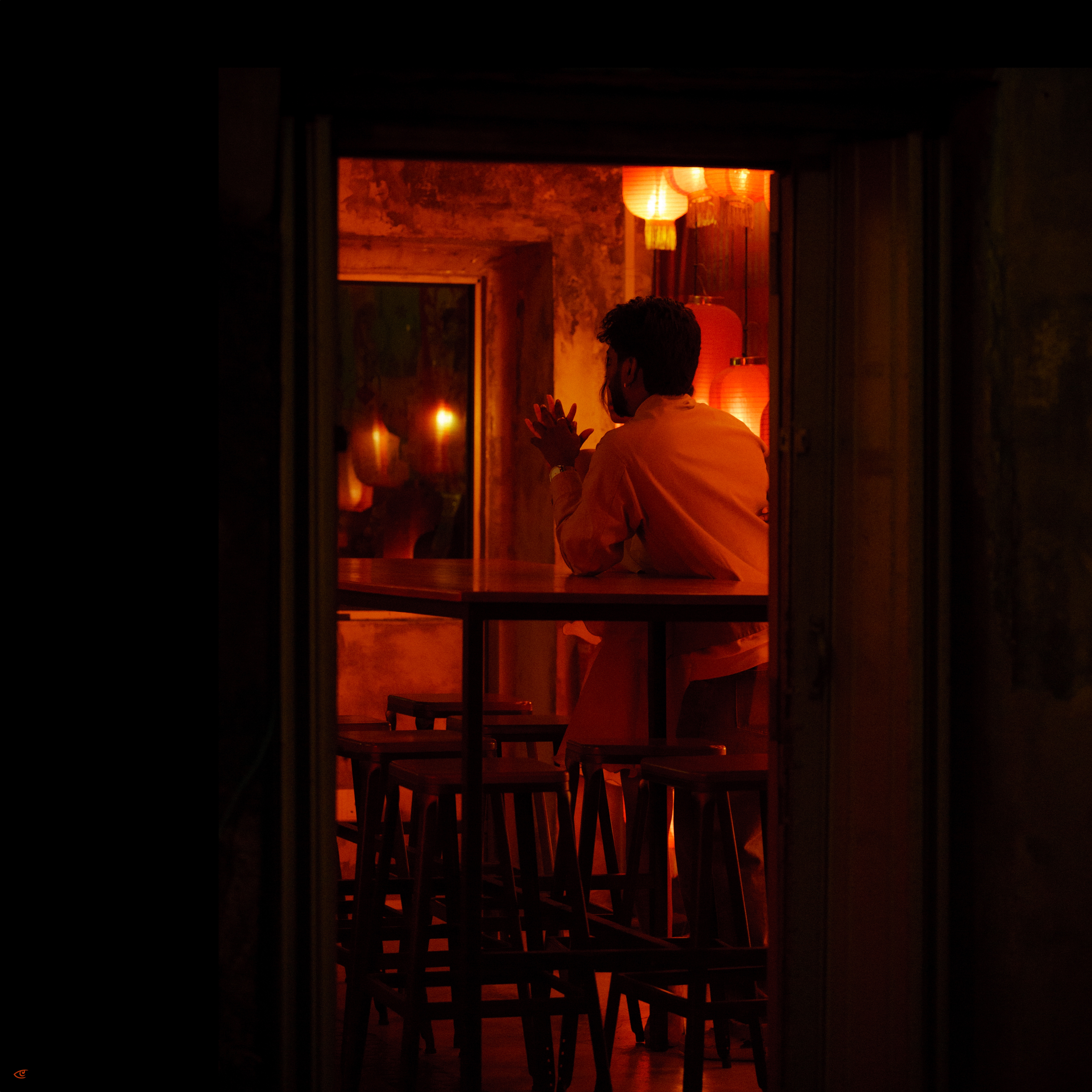 Person seated at a bar counter, seen in profile through a dark doorway, with hands near their face; warm orange lantern lights and a wall mirror behind them, and tall stools lined up in the foreground.
