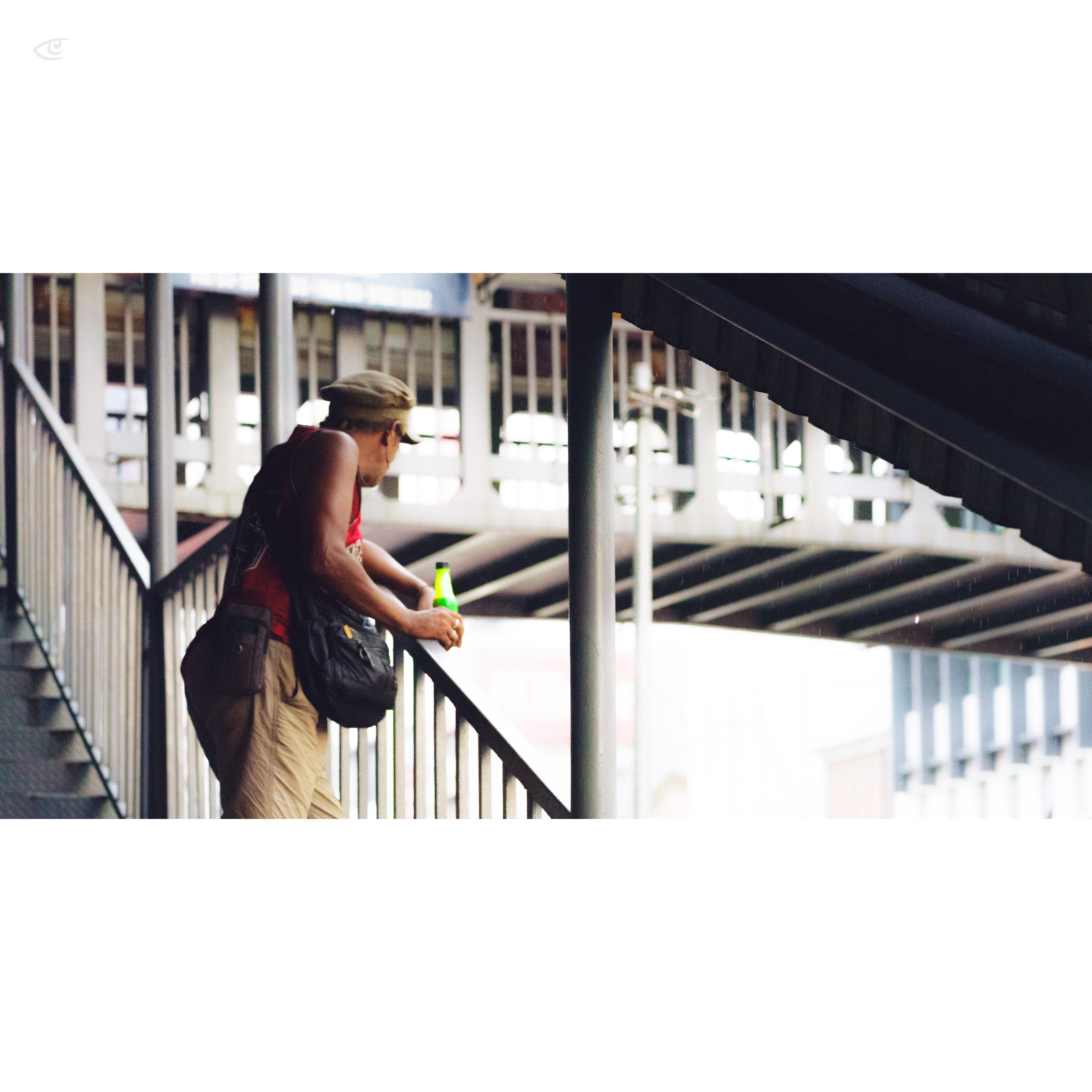 Person in a red tank top and khaki pants leans on a metal railing under an elevated structure, holding a green bottle, with stairs and steel beams in the background.