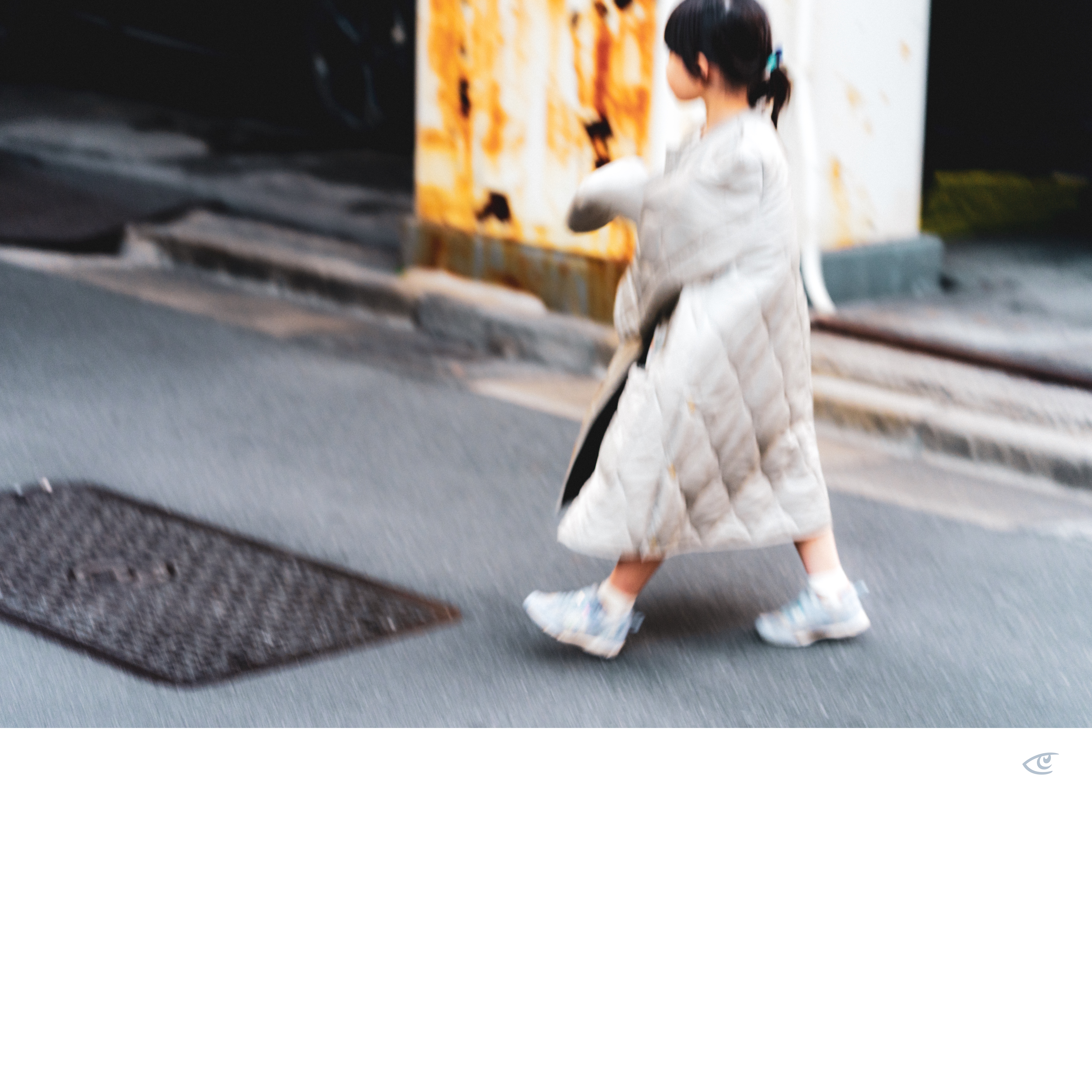 Blurred side view of a child with a ponytail walking on a street, wearing a long quilted coat and sneakers, with a curb and a rust-stained wall in the background.