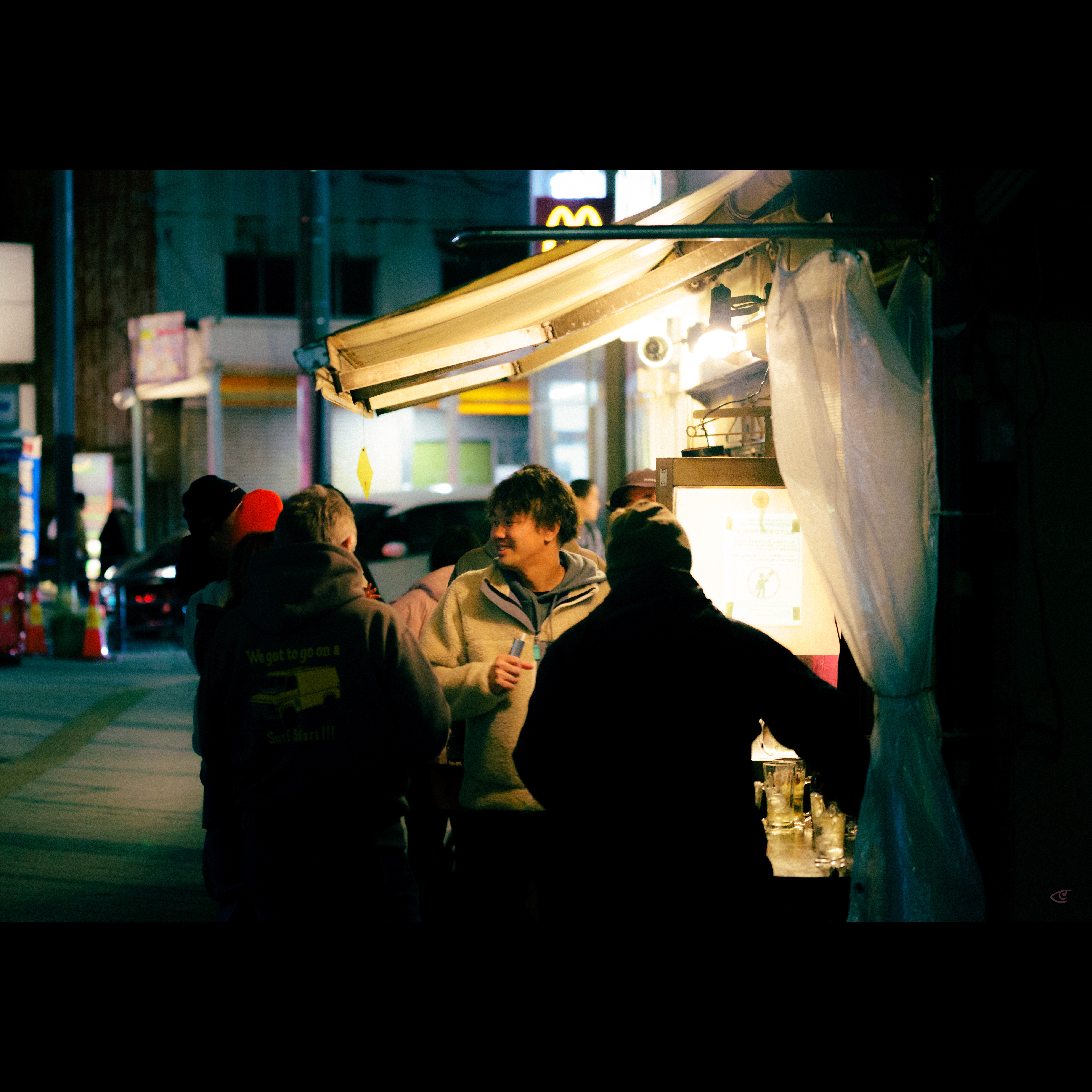 Group of people gathered at a small street food stall at night, lit by bright hanging bulbs under an awning; one person in a light jacket holds a drink while others stand near a counter with cups, with a dim city street and parked cars in the background.