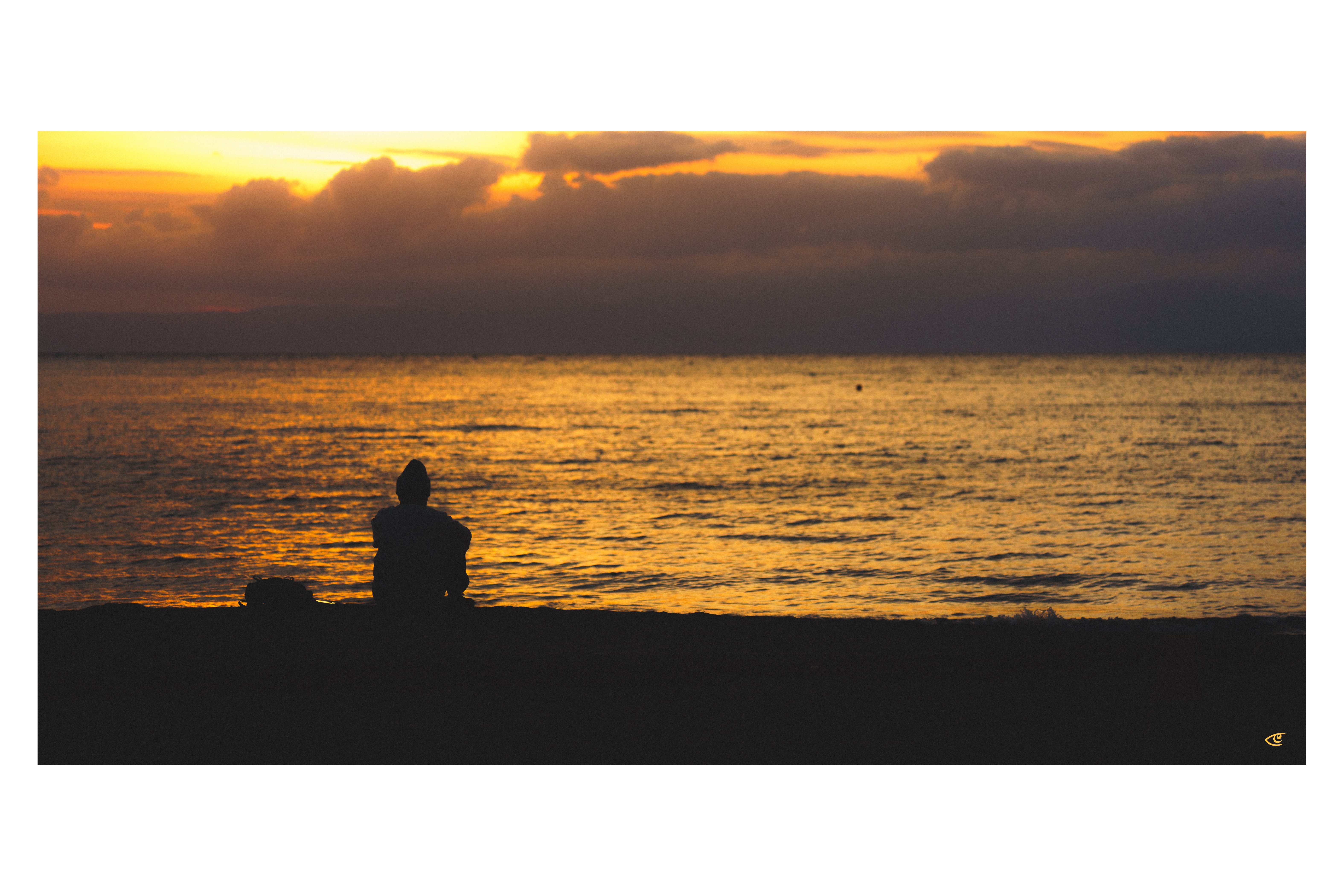Silhouette of a person sitting on a dark shoreline facing the ocean, with golden sunset light reflecting on the water and thick clouds across the horizon.