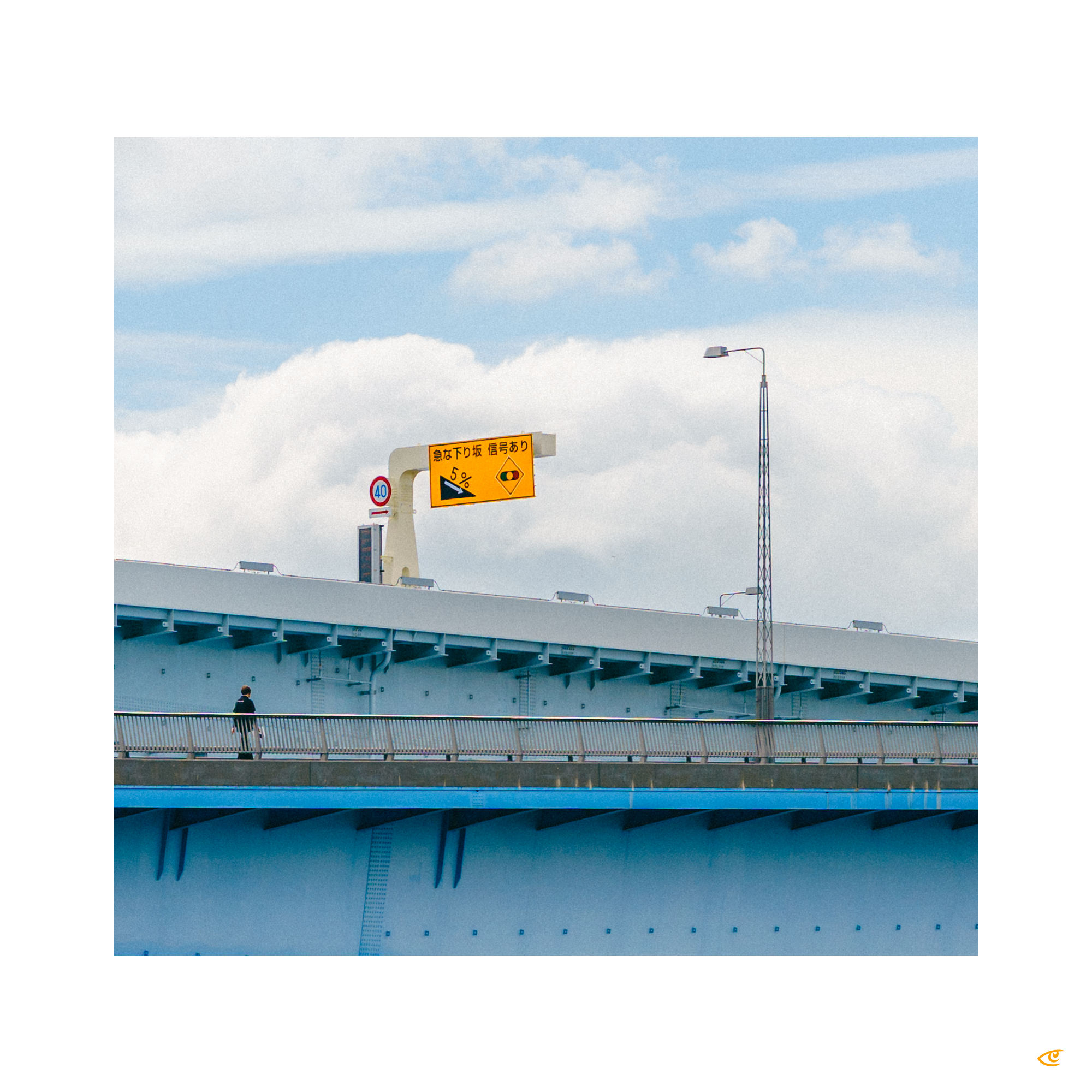 Pedestrian walkway on a large blue bridge with a lone person walking along the railing; overhead yellow road sign with Japanese text and a 40 speed limit sign against a cloudy sky.