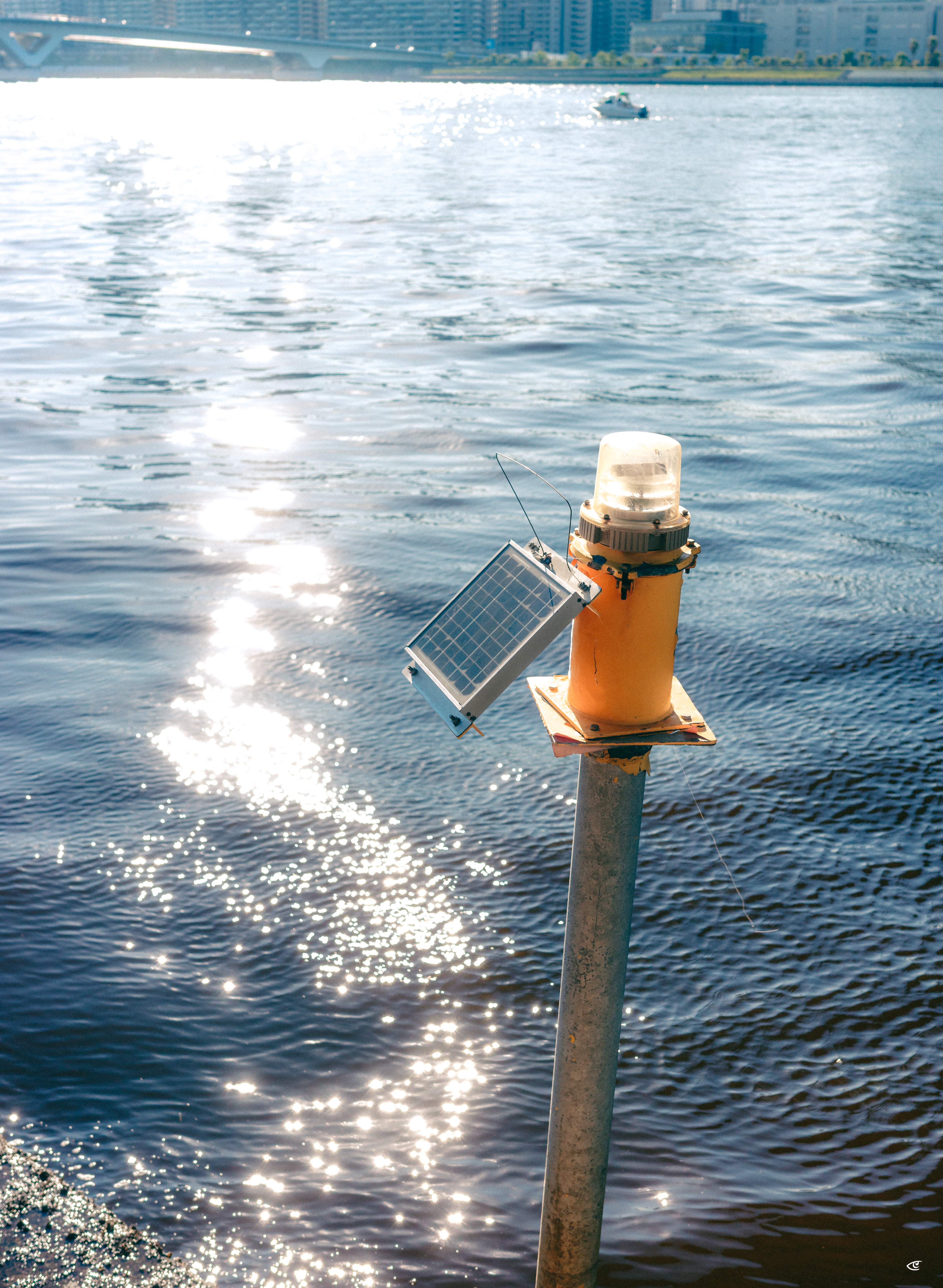 Yellow navigation beacon with a clear lens and a small solar panel mounted on a metal pole beside a rippling river, with sun glare on the water and a distant bridge and city buildings in the background.