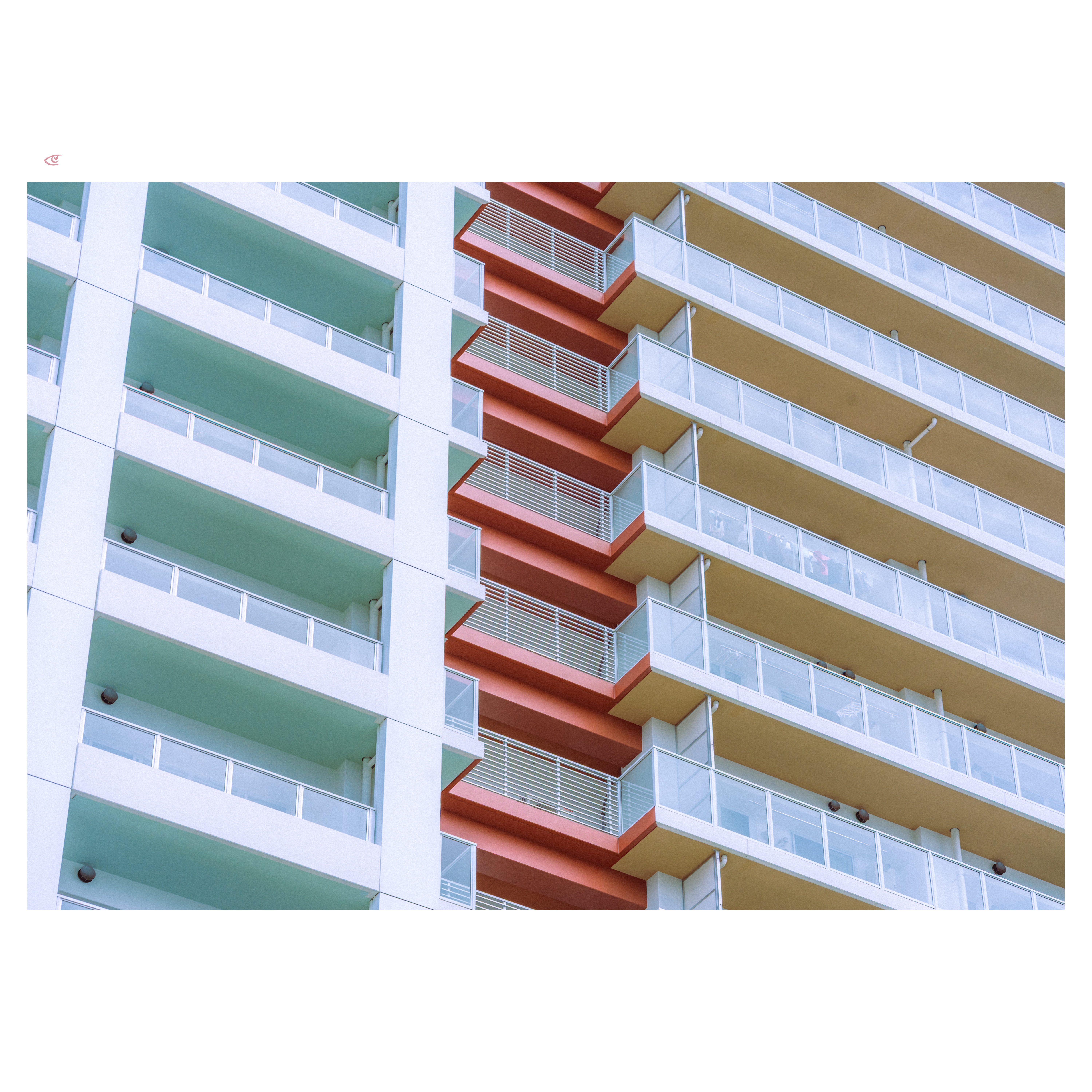 Diagonal close-up of a modern apartment building facade with stacked balconies; mint-green balconies on the left, beige balconies on the right, separated by a red central column with metal railings and glass panels.